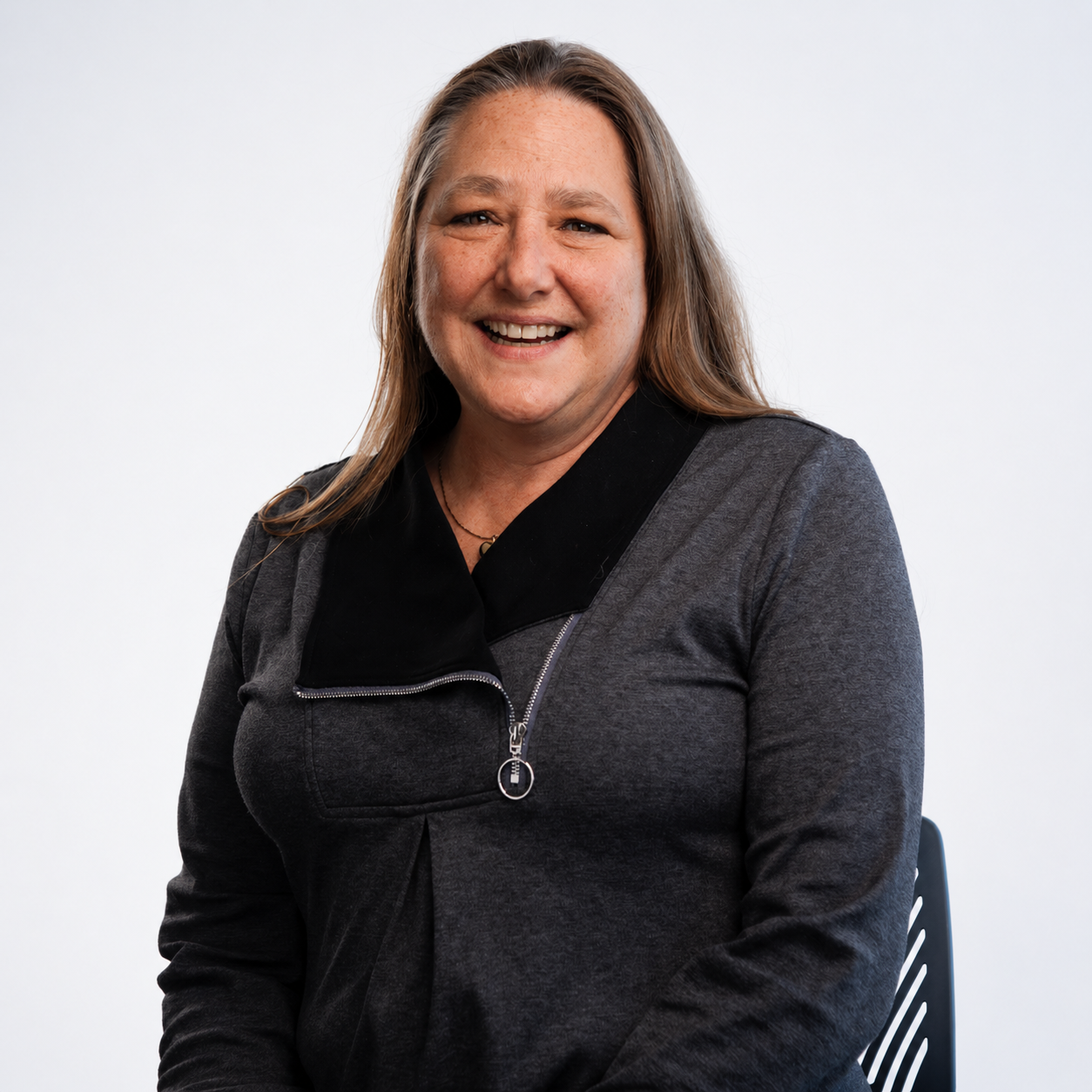 Meet our education administrator, Kim Bacon. A woman smiling outdoors on a cloudy day, standing on a grassy area with trees and a landscape in the background, wearing a brown cardigan and a beaded necklace.