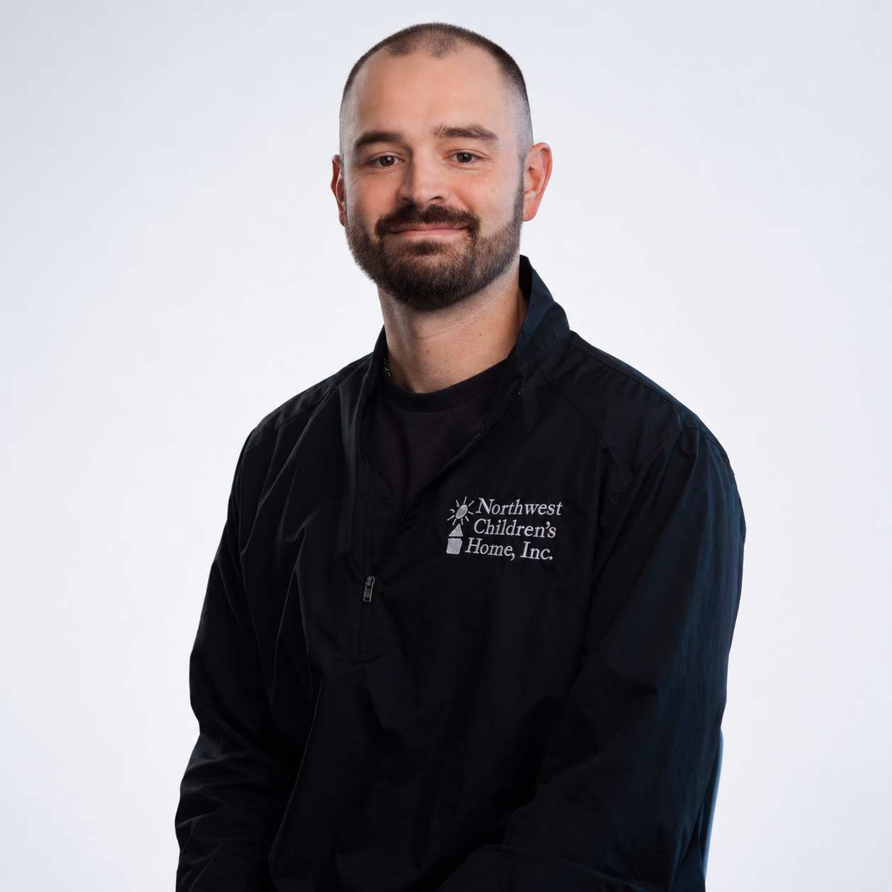 Meet our Residential Program Administrator, Logan Woolery. A man kneeling on a grassy area outdoors with a brown dog. The man is wearing a black jacket with a logo and text that says 'Northwest Children's Home, Inc.', a black cap, and jeans.
