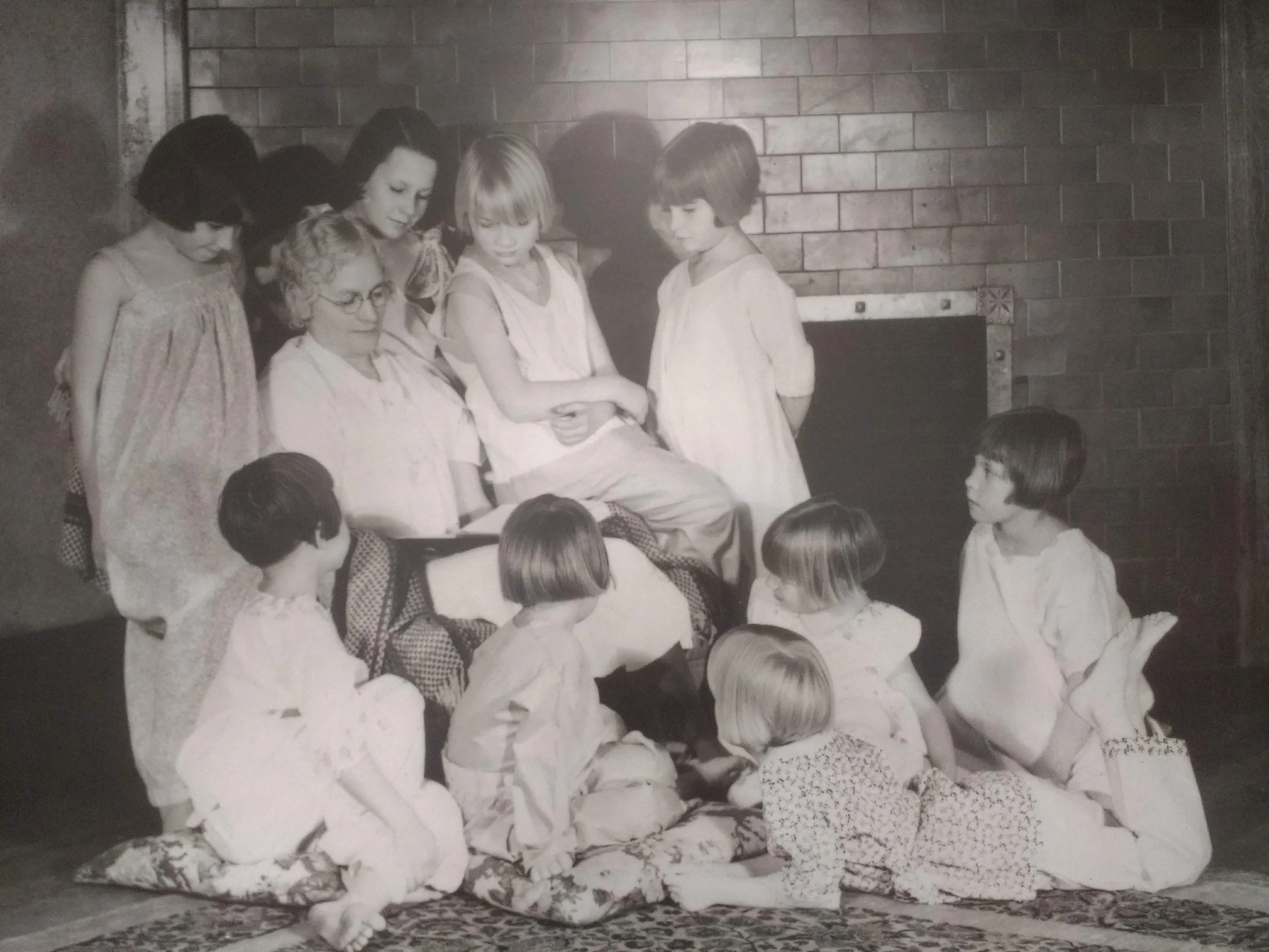 A group of children and an adult woman gathered around a seated woman, with some children sitting on the floor and others standing, engaged in a storytelling or performance in front of a brick wall.