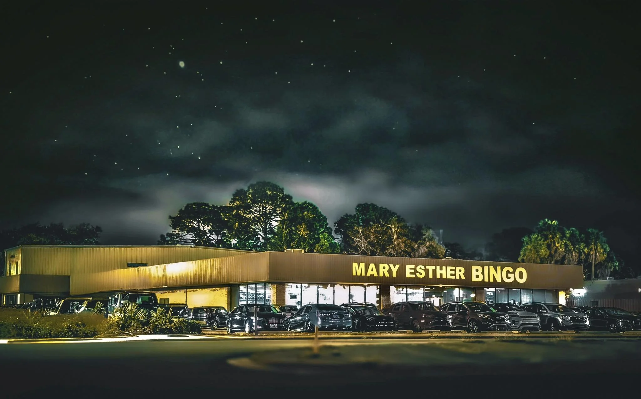 Night view of a bingo hall named 'Mary Esther Bingo' with cars parked in front and trees behind, under a starry sky with clouds.