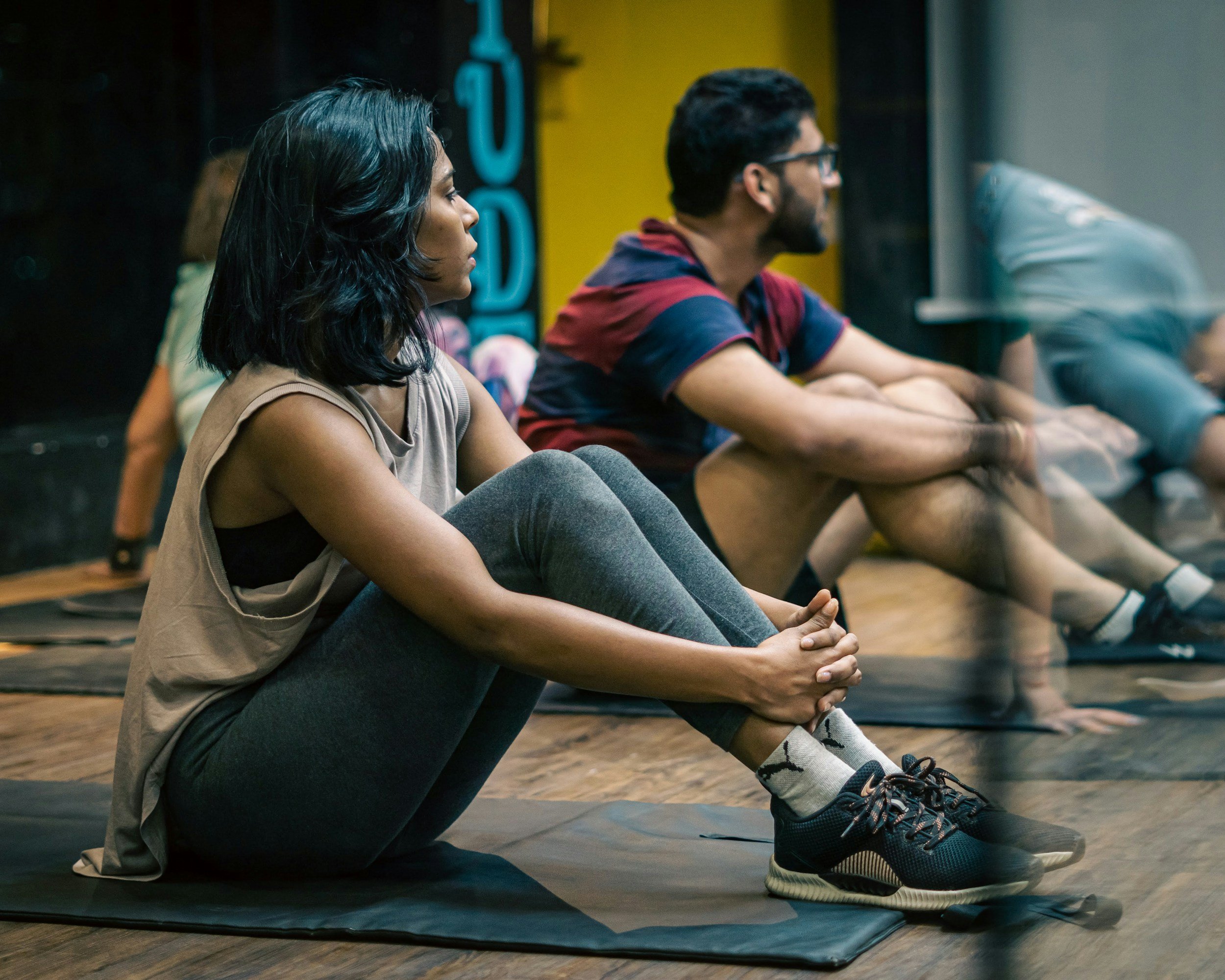 A group of people sitting on yoga mats in a calming indoor space, with two individuals in the foreground, a woman with black hair and a man with glasses and a beard, both seated cross-legged and looking straight ahead, possibly in a meditation or yoga class.