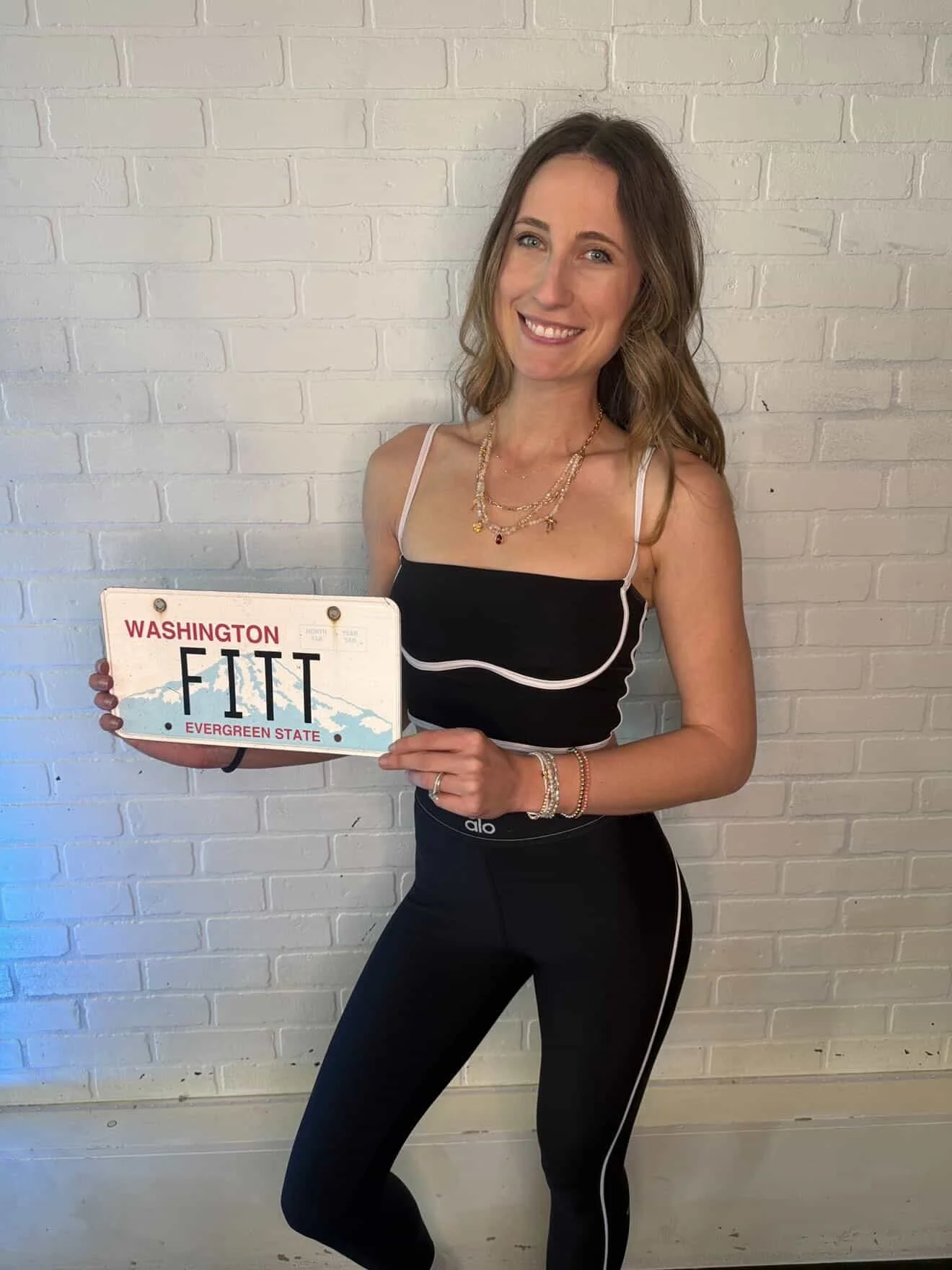 A woman with light skin, wavy brown hair, and a big smile, standing against a white brick wall, holding a license plate that reads 'WASHINGTON FIT.' She is wearing a black athletic top with white straps, black leggings with white side stripes, multiple gold necklaces, and bracelets.