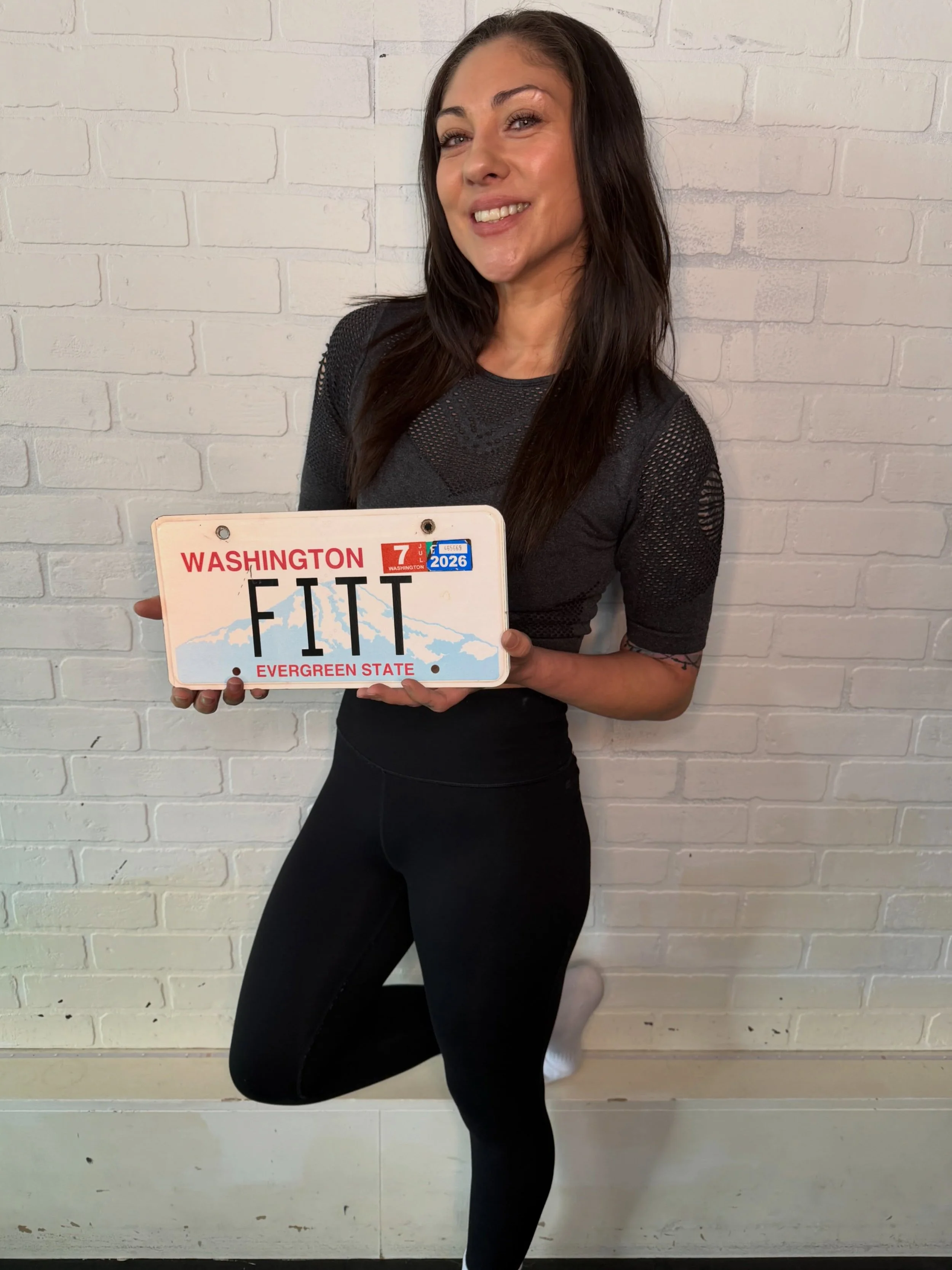 A woman with long dark hair wearing a black athletic outfit, holding a Washington license plate with the words "FIT" and "Evergreen State" showing, standing against a white brick wall.