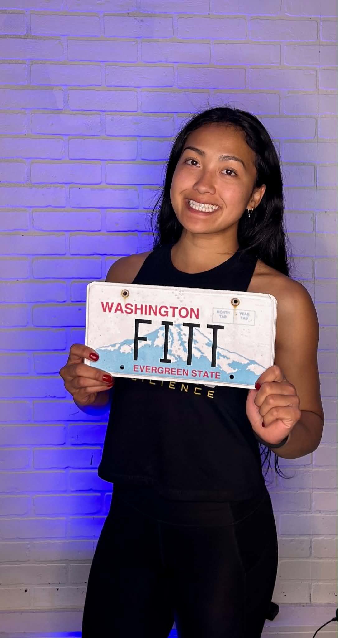 A woman with long black hair smiling and holding a Washington license plate that says 'FITT' and has a mountain graphic, standing against a white brick wall with purple lighting.