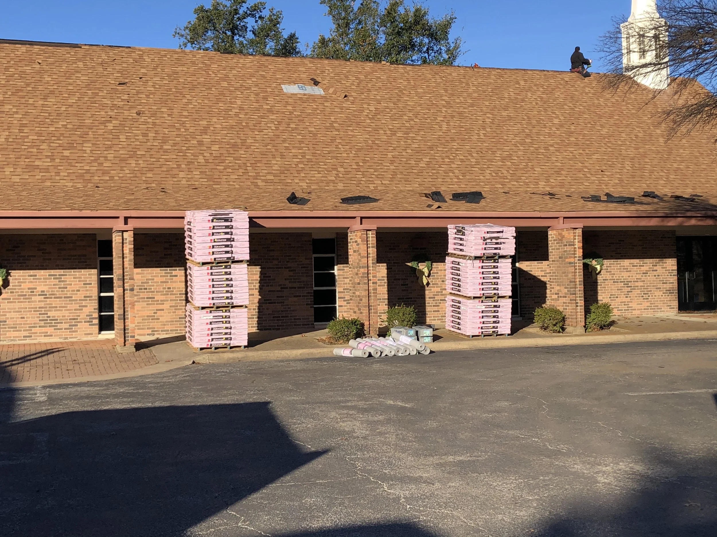 Workers are installing roofing materials on a house under construction, with plywood and roofing shingles visible.