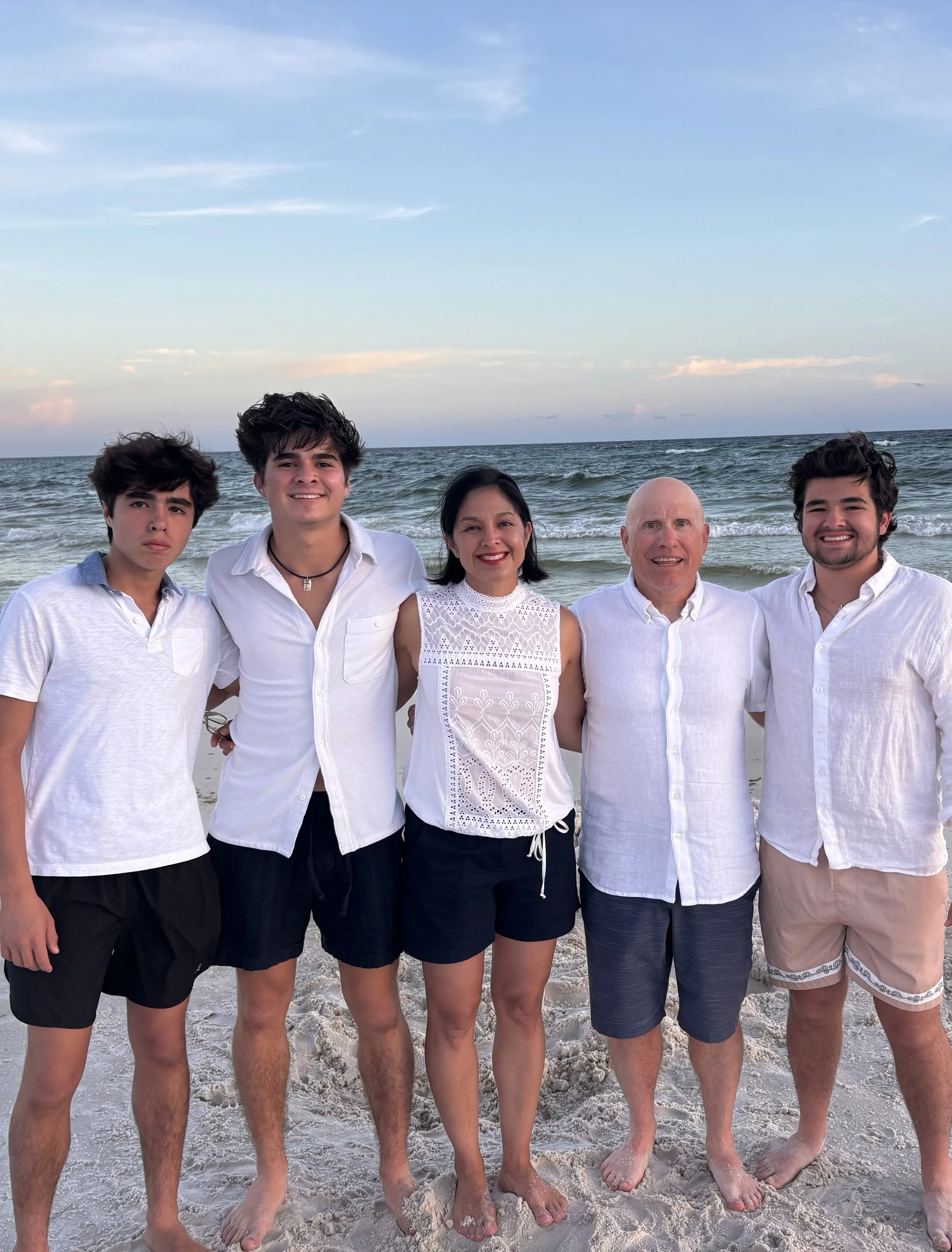 Family standing on the beach with the ocean and sky in the background, all dressed in white and light-colored clothing.