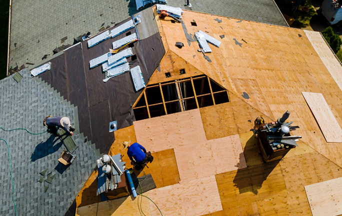 Workers are installing roofing materials on a house under construction, with plywood and roofing shingles visible.