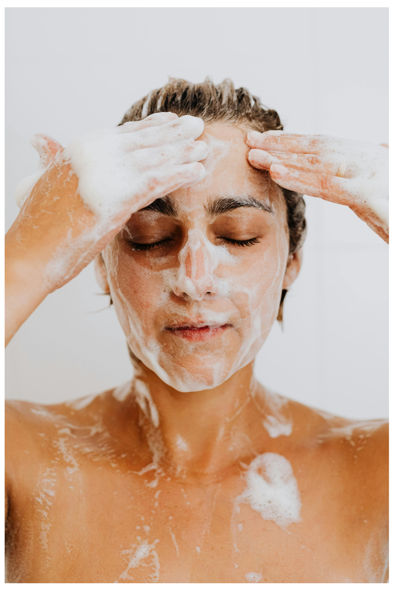 A person taking a shower, with soap and foam on their face and upper body, eyes closed.