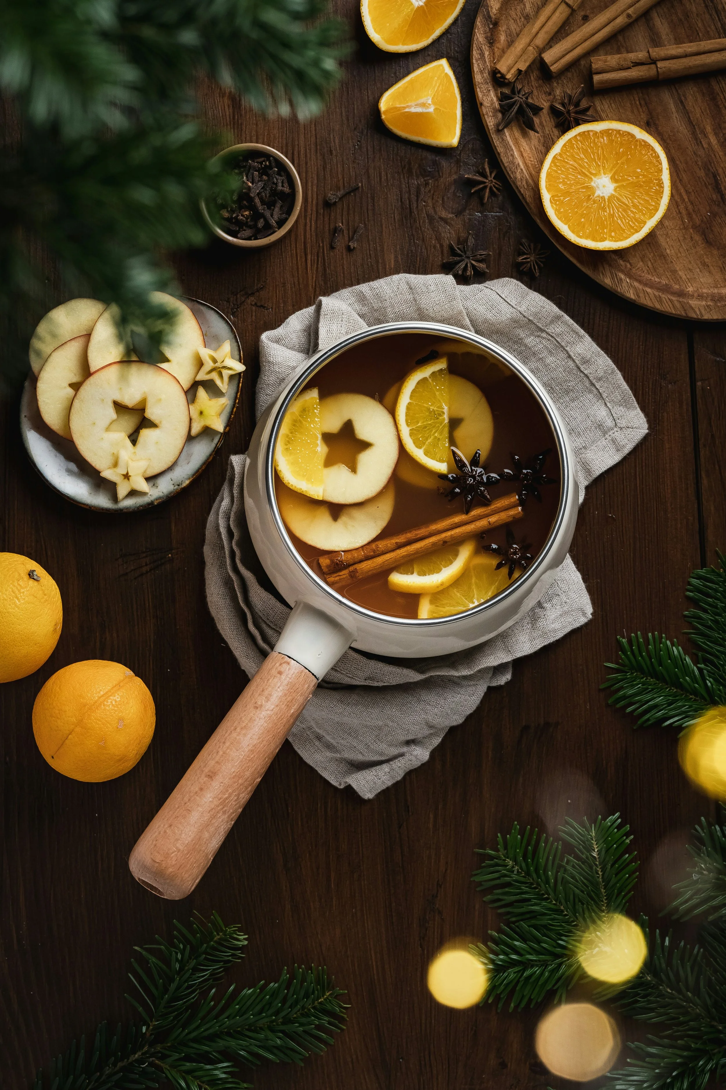 A saucepan of apple cider with lemon slices, apple slices, star anise, and cinnamon sticks on a wooden table surrounded by lemons, apple slices, and holiday decorations.