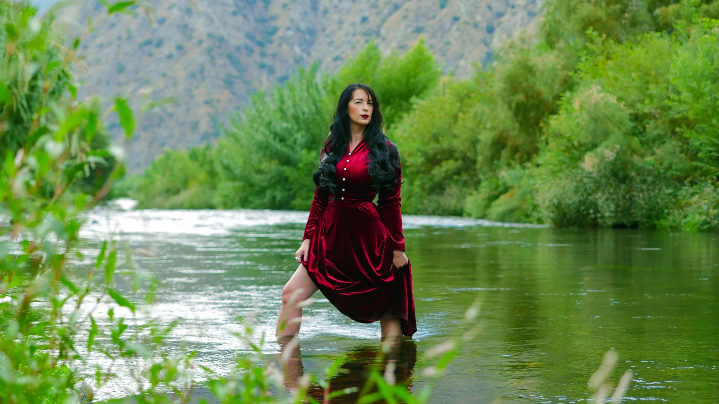 Jasmine Maya Royce in Los Angeles National Park in a velvet red dress standing in a river surrounded by lush green trees and mountains in the background.