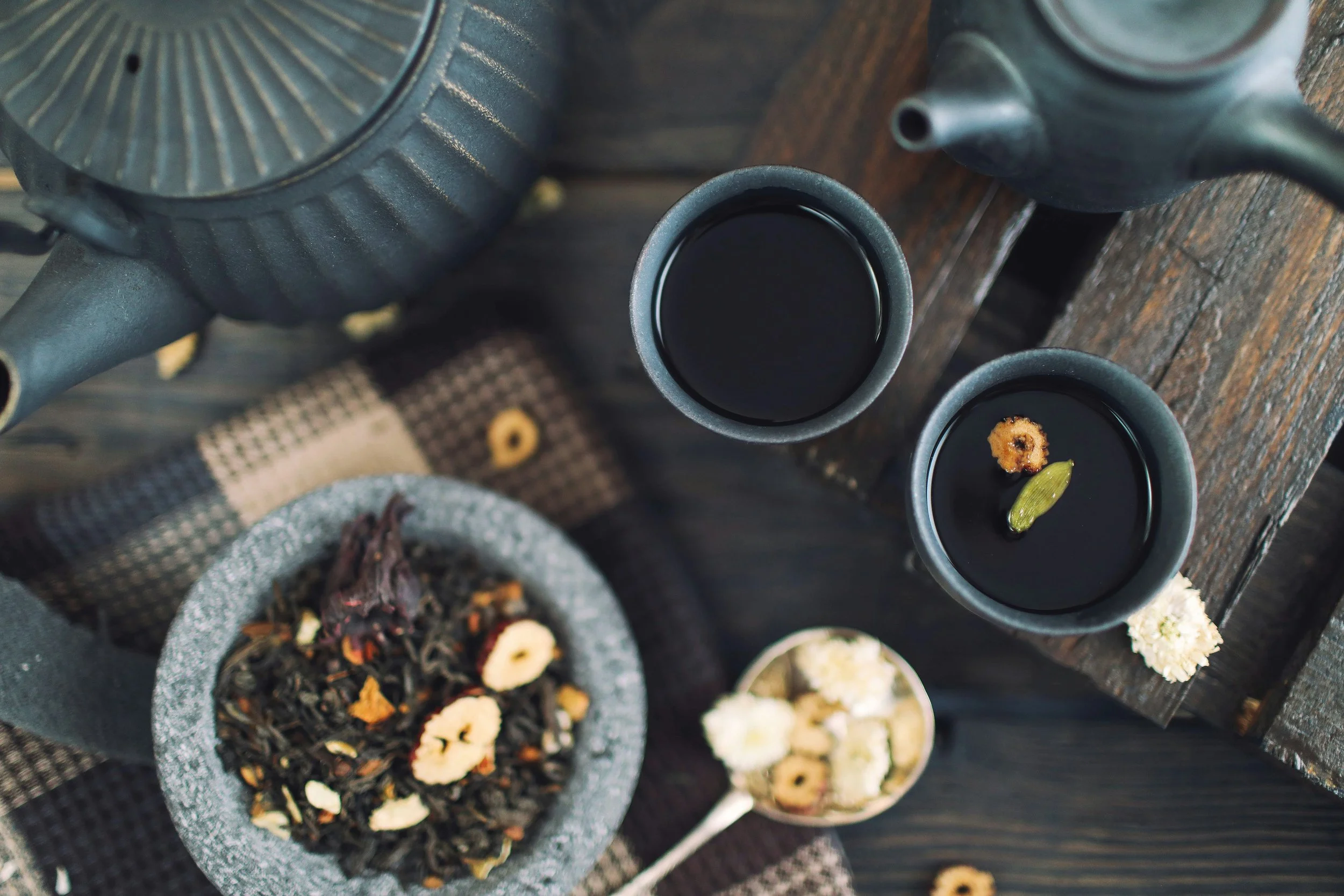 Overhead view of two cups of black tea with floating flowers and a piece of green cardamom, placed on a wooden surface. A teapot, a bowl of loose tea leaves with dried flowers and banana chips, and a small spoon with dried flowers are also visible.