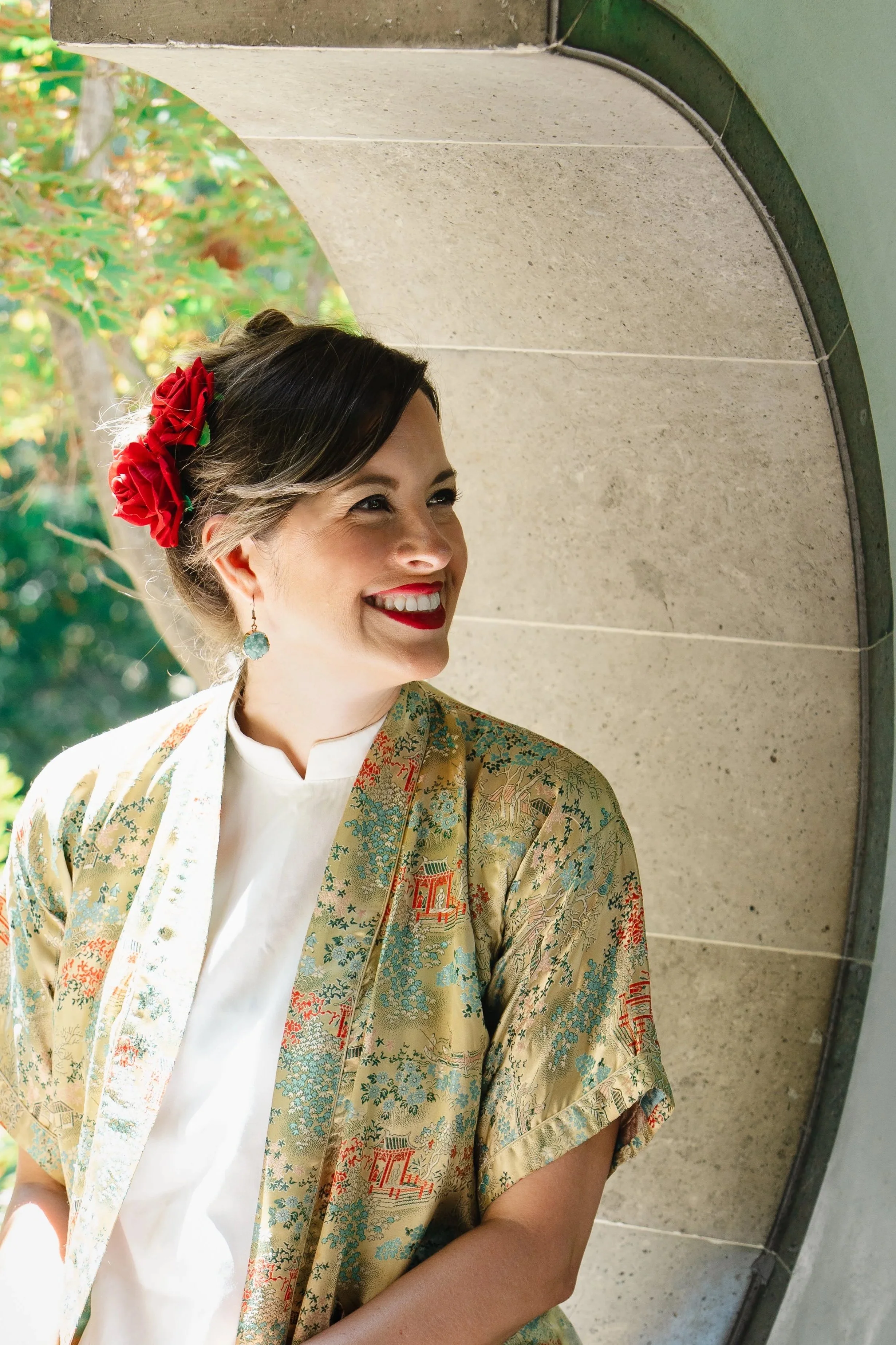 Jasmine Maya Royce styled with dark hair and red lipstick, smiling while looking to her right, wearing a colorful kimono and red flower hair accessories, standing under an arched stone structure with green foliage in the background.