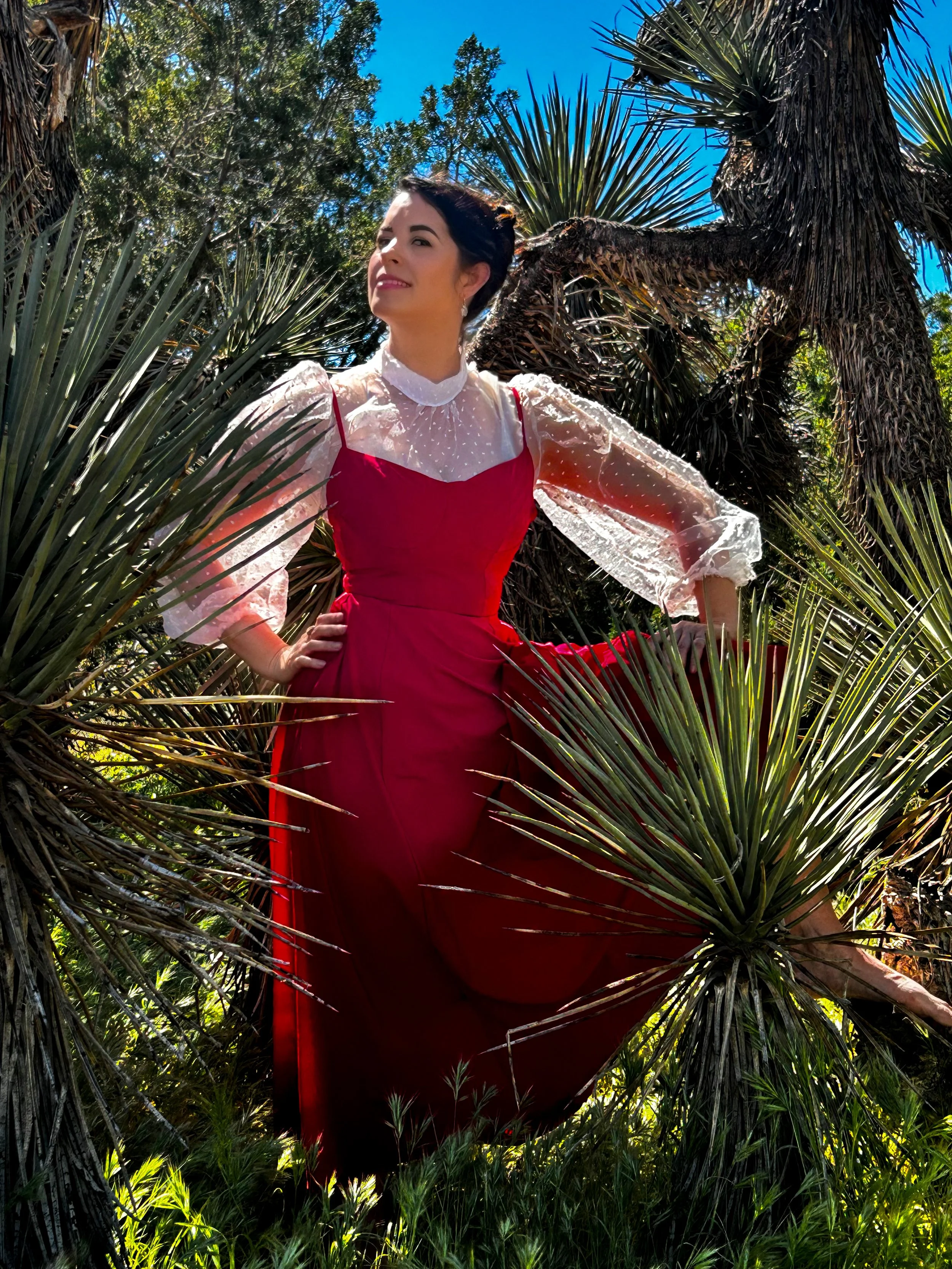 A woman wearing a red dress with puffy white sleeves standing among desert plants, with a bright blue sky in the background.