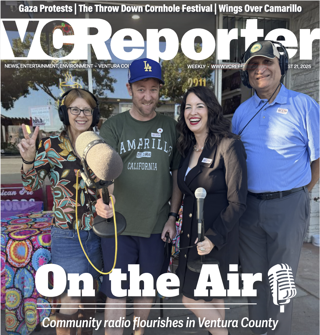 Four board members including Jasmine Maya Royce standing outside at a community radio station event, holding microphones, with one woman making a peace sign, all smiling. The scene is lively with a colorful tablecloth