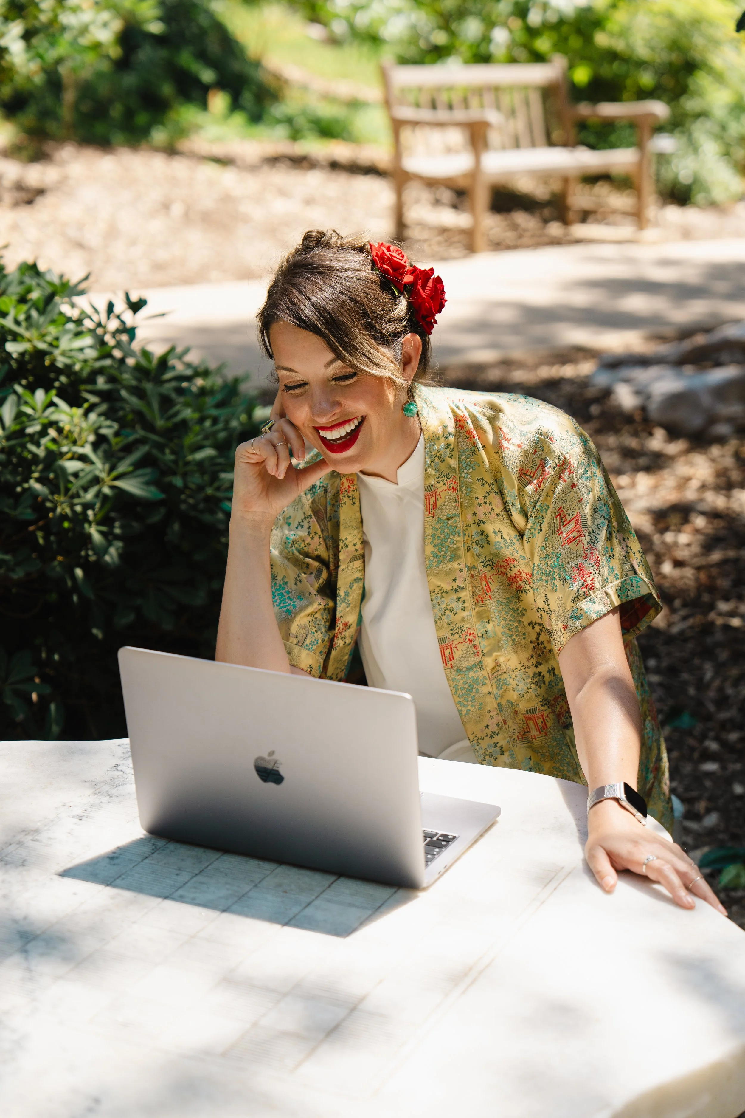 Jasmine Maya Royce styled with dark hair styled with red flowers, wearing a colorful kimono-style jacket, sits at a table outdoors, smiling while looking at her laptop.