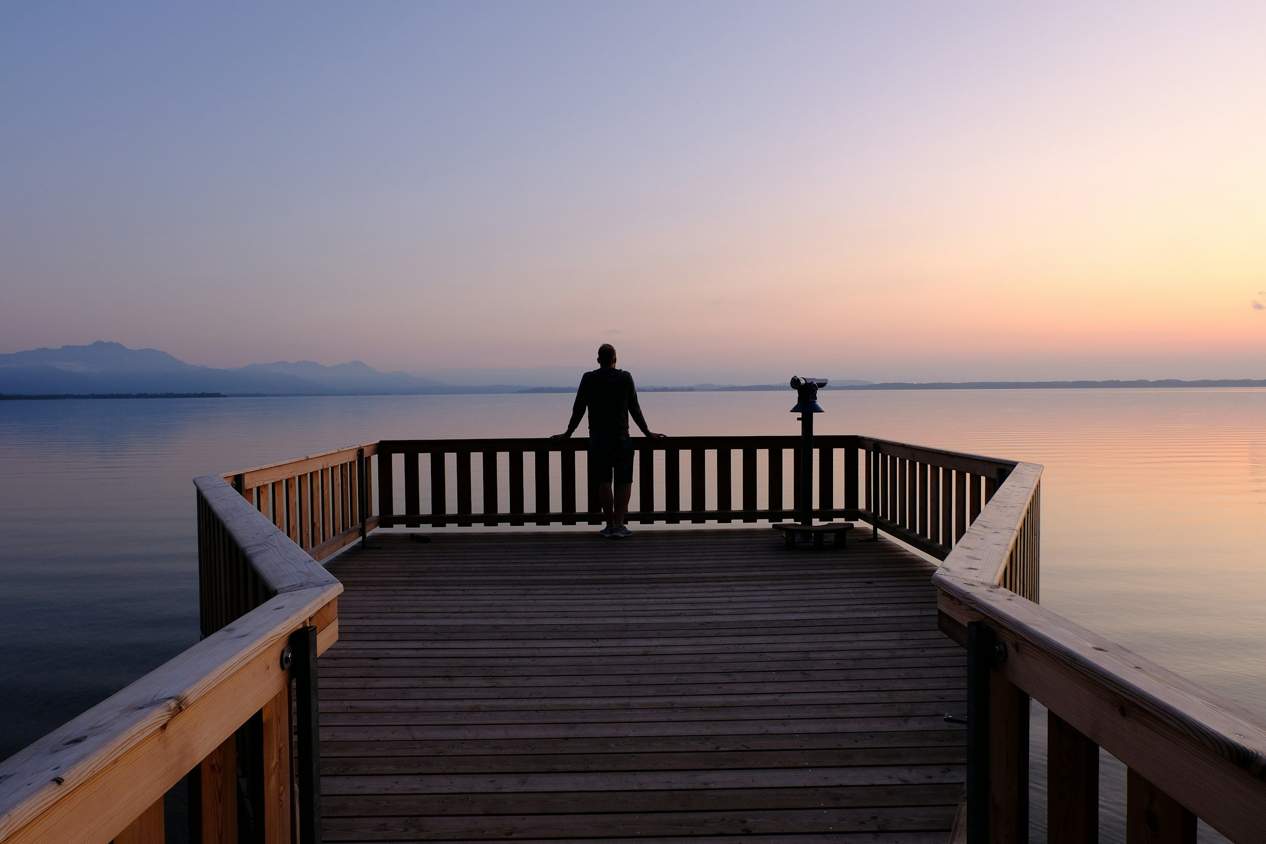 A person standing on a wooden pier, looking out over calm water at sunset with a faint mountain range in the distance.