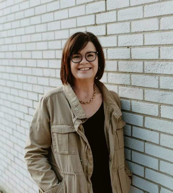 A woman with short dark hair and glasses standing against a white brick wall, smiling, wearing a tan jacket over a black top and a gold chain necklace.