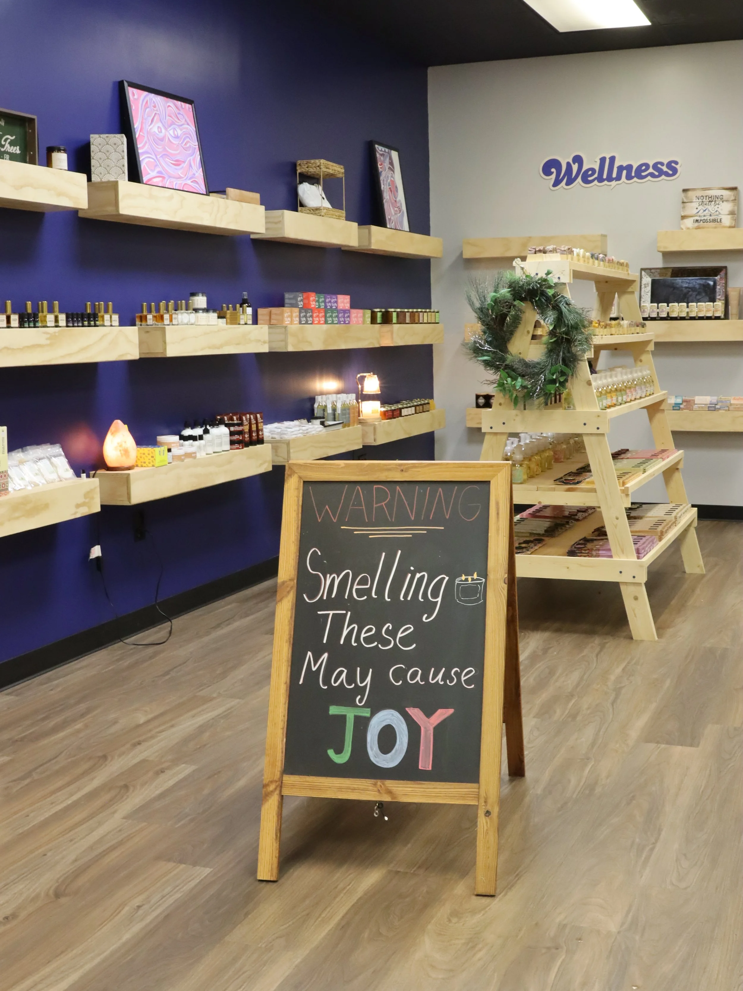 A retail store with wooden shelves on a blue wall displaying small bottles and products, a chalkboard sign warning about fragrances causing joy, and a decorative wreath on a wooden display ladder, with the word 'Wellness' on the wall.
