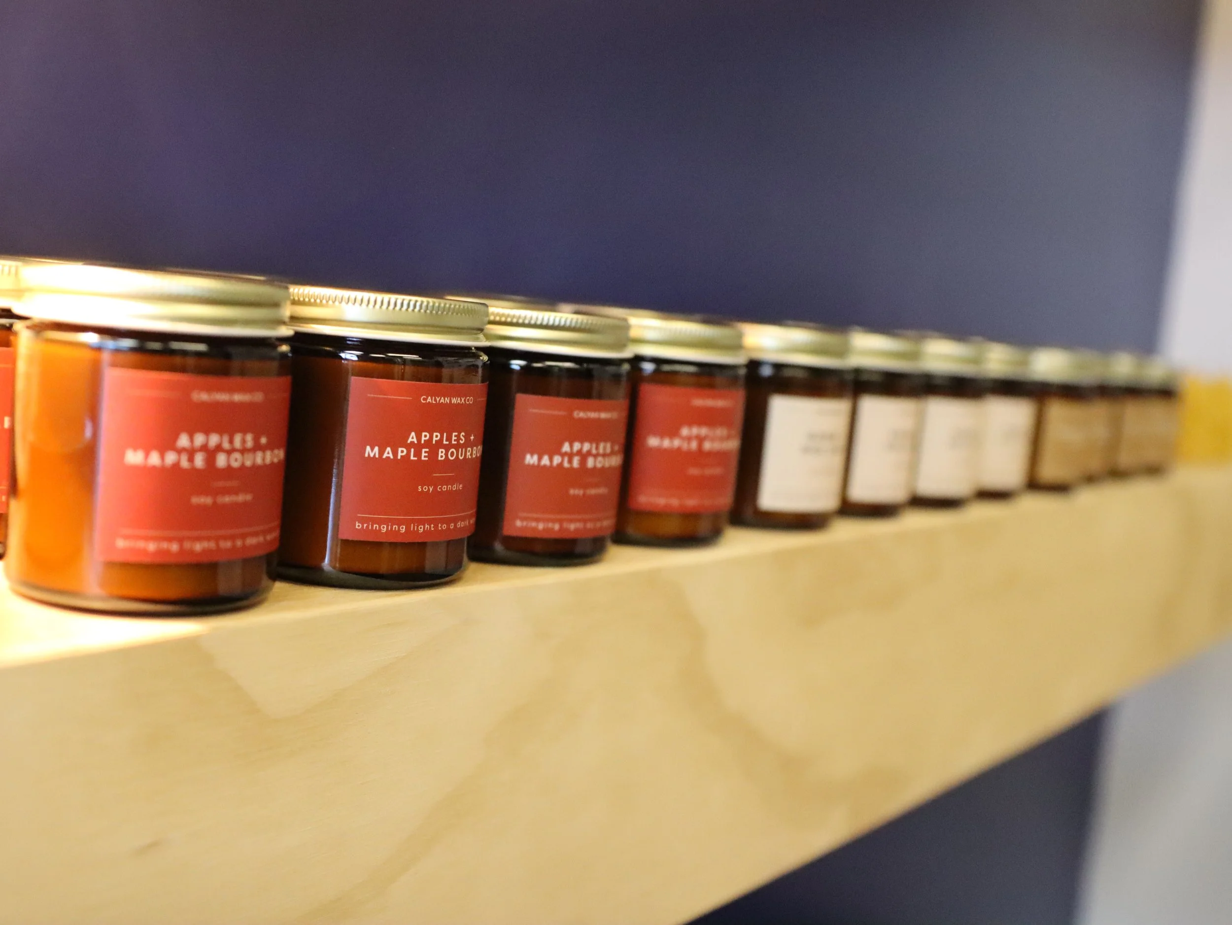 A row of scented candles on a wooden shelf, with labels reading 'Apples + Maple Bourbon' and a dark background.