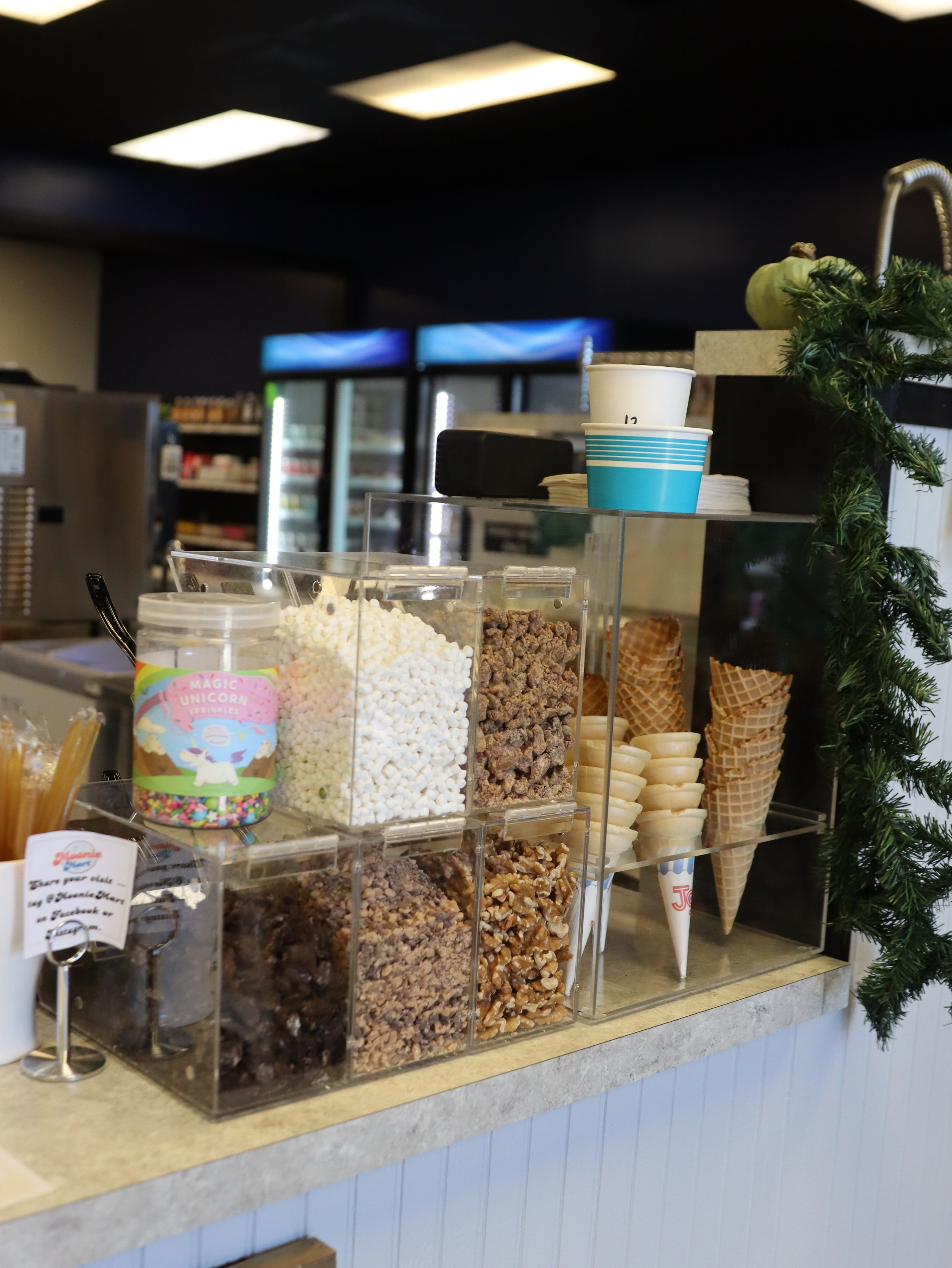 Display of various toppings and ice cream cones at an ice cream shop counter.