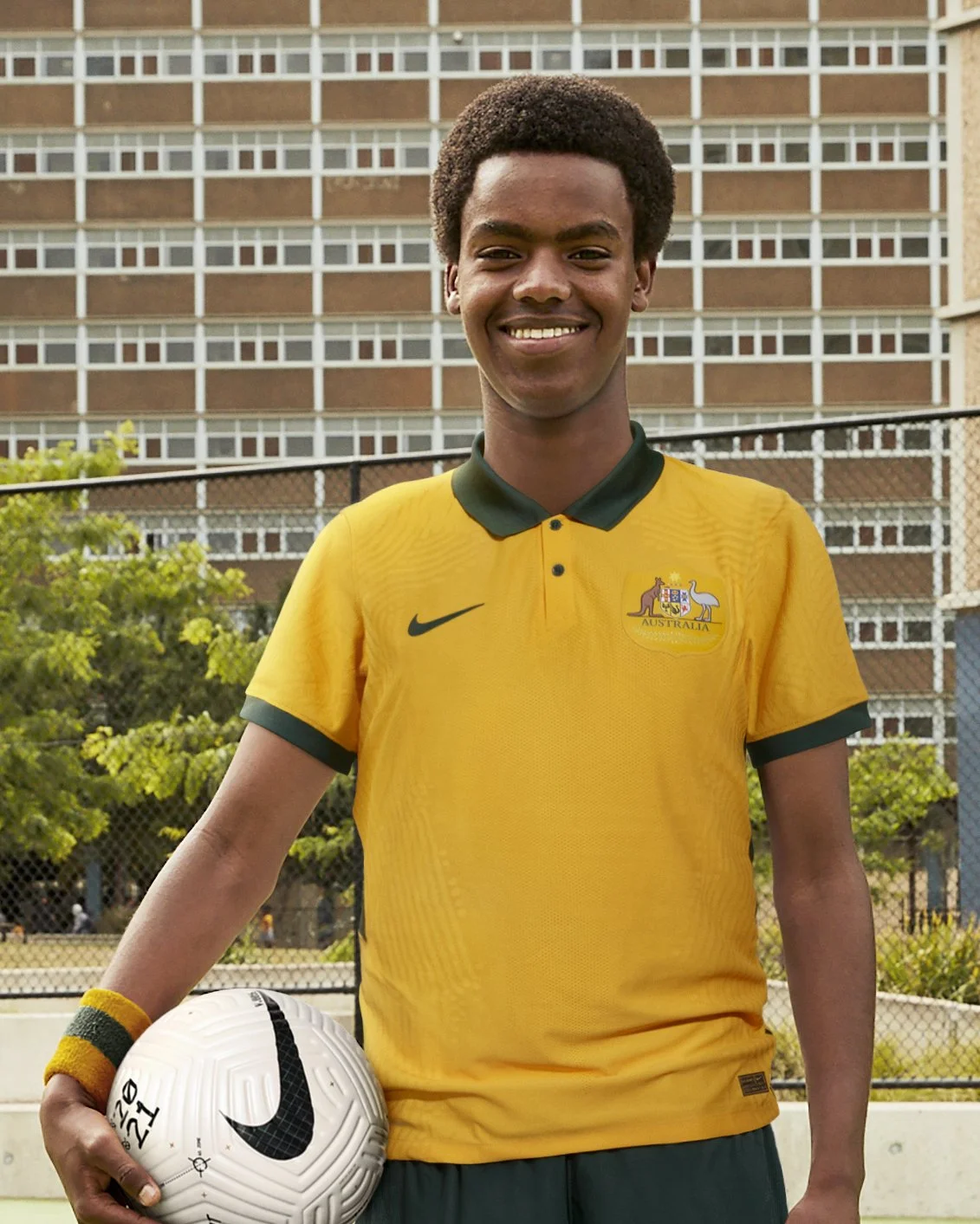 Young man in yellow Australia soccer jersey holding a white soccer ball with black Nike logo outdoors on a sports court, with a multi-story building and trees in background.