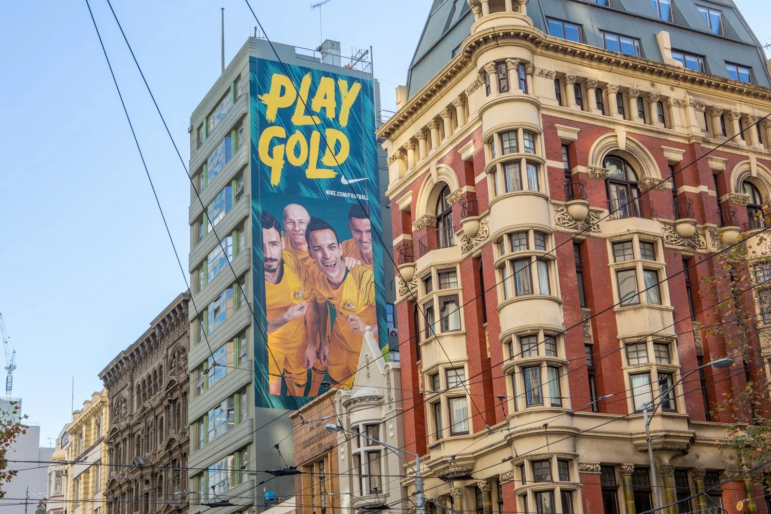 A large billboard on a tall building featuring four men in yellow sports uniforms, smiling and holding medals, with the text "Play Gold" in bold yellow letters. The background of the billboard has a blue abstract design, and the website nike.com. The billboard is on an urban street with ornate older buildings and various wires crossing in the foreground.