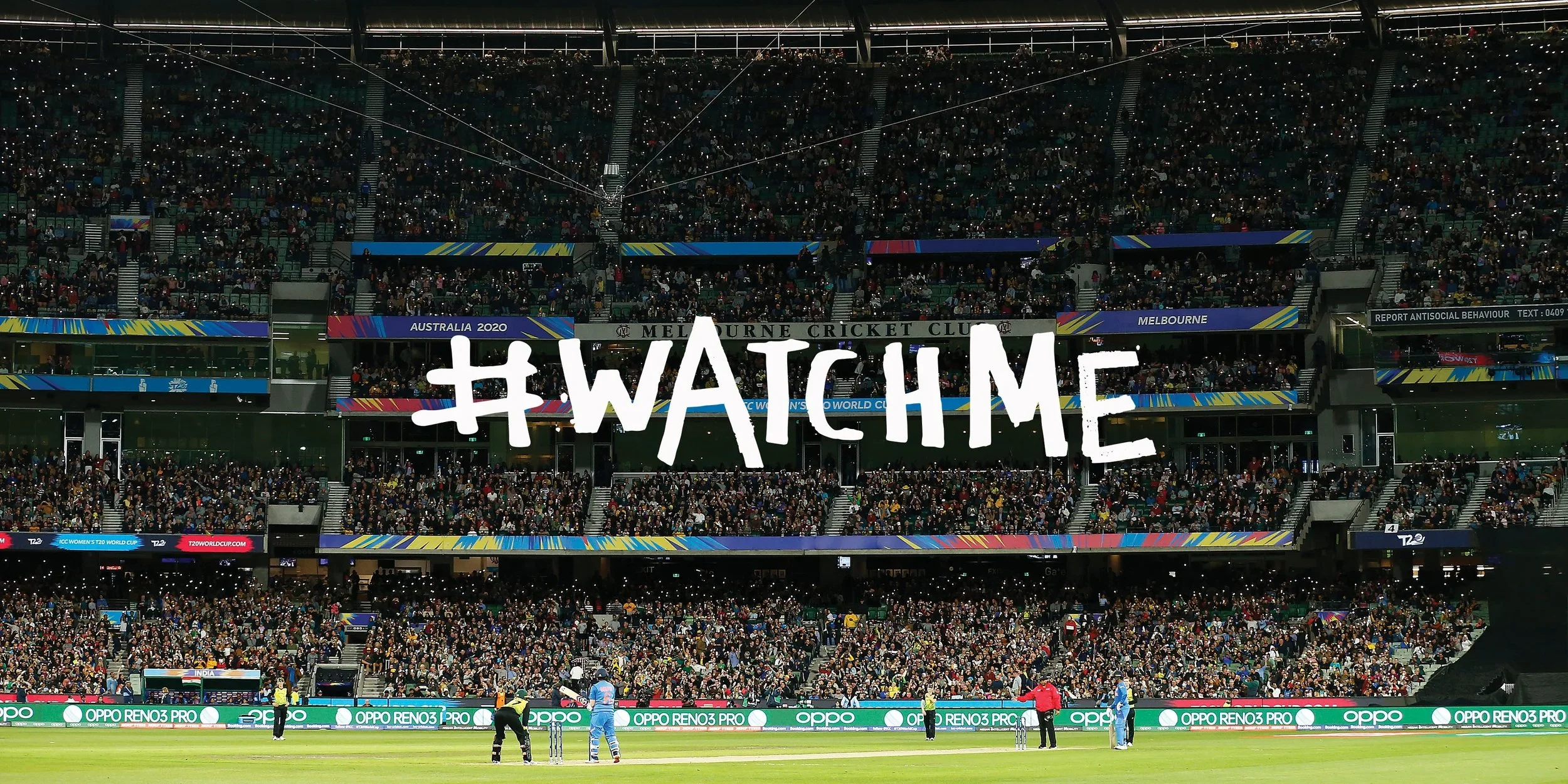 View of a cricket stadium filled with crowd during a match with the hashtag #WATCHME and colorful banners, with players on the pitch.