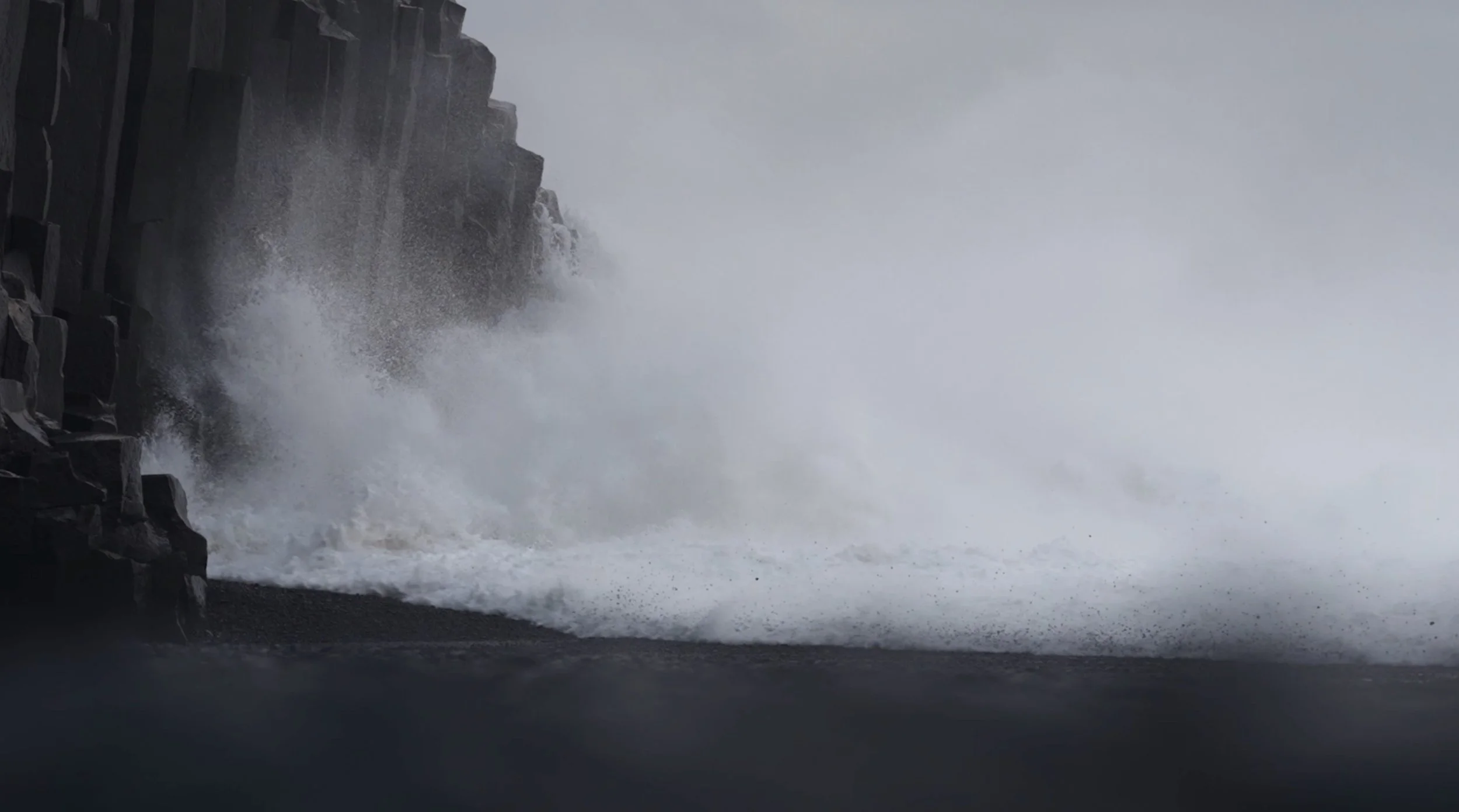 Ocean waves crashing against dark rocky cliffs during a stormy day with gray sky.
