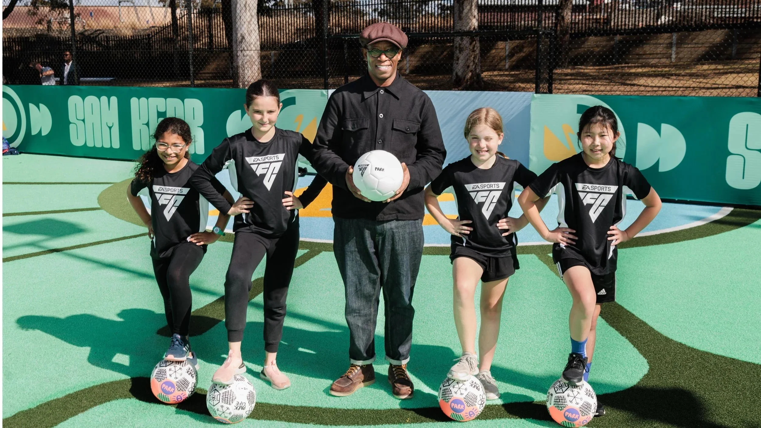 A man in a black jacket and cap holding a soccer ball standing on an artificial turf soccer field with four young girls in matching black sports uniforms, each with a soccer ball at their feet, all smiling and posing for a group photo.