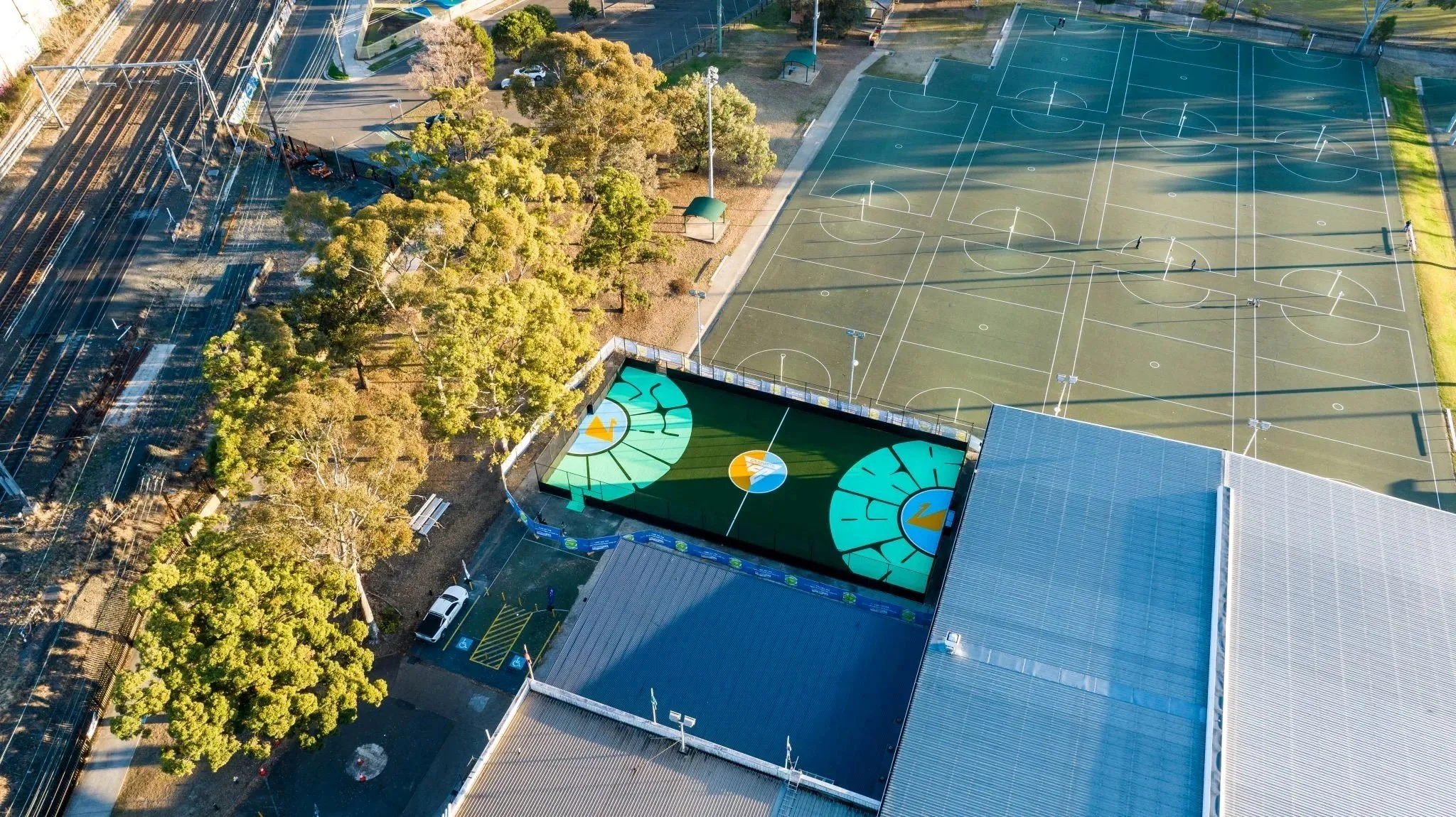 An outdoor sports complex with multiple basketball courts, a small parking lot with a few vehicles, trees lining the area, and a distinctive rooftop maze with geometric shapes and vibrant colors.