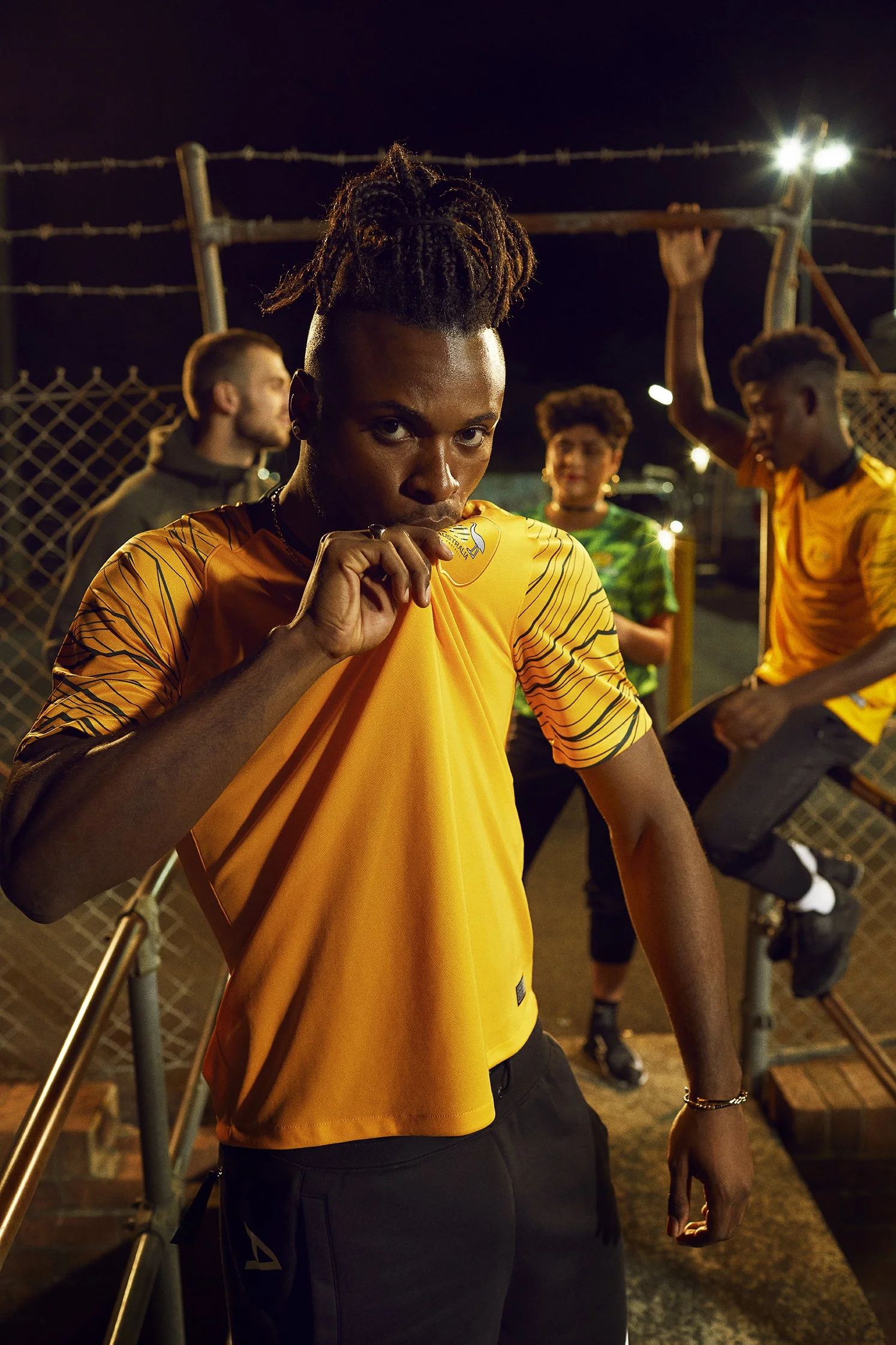 A woman in a yellow sports jersey poses confidently in the foreground at night, with three other young people in sporty attire in the background near a chain-link fence, illuminated by artificial lights.