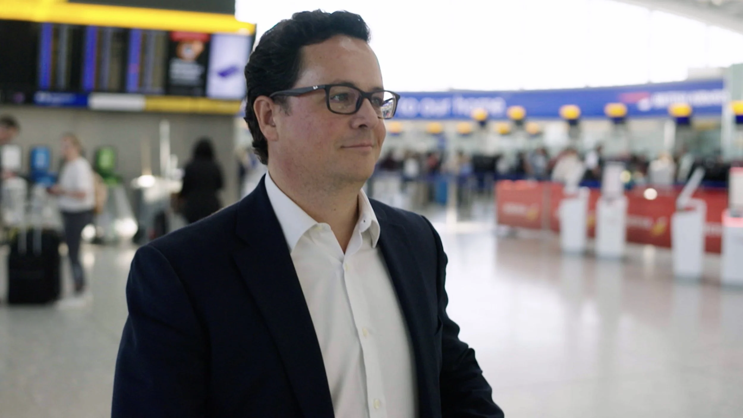 A man with dark curly hair, wearing a dark suit and glasses, standing in an airport terminal with check-in counters and electronic displays in the background.