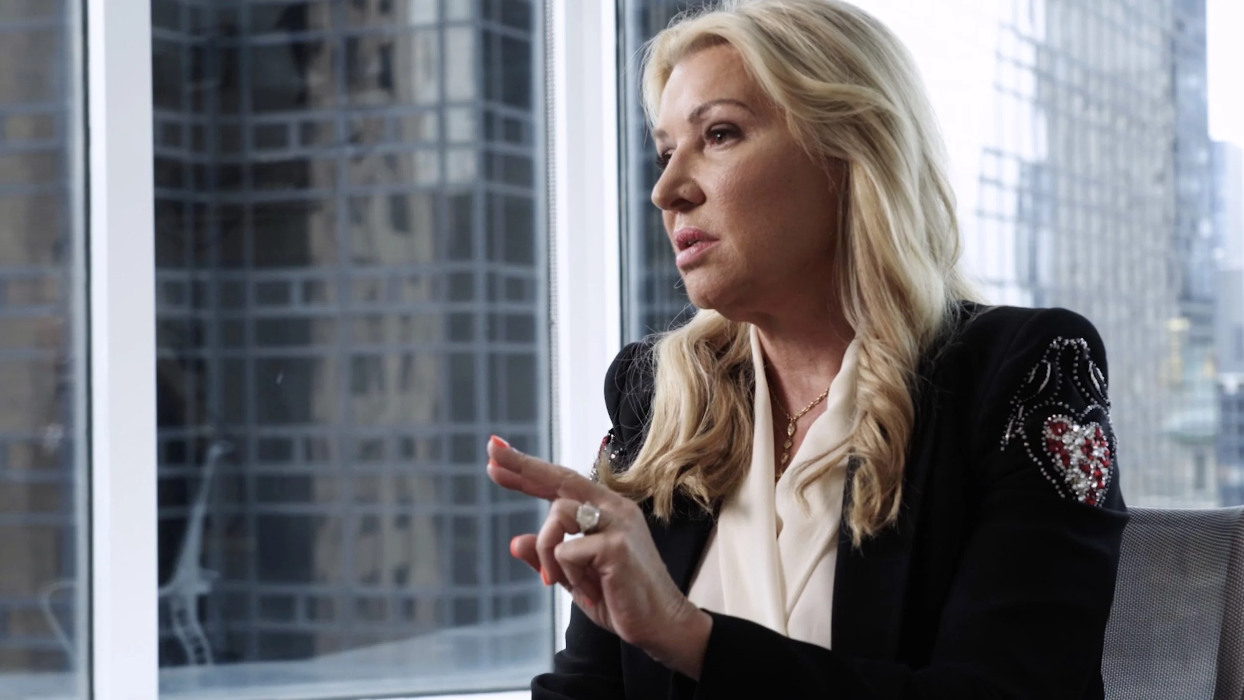A woman with blonde wavy hair, wearing a black blazer with decorative embroidery on the shoulders and a white blouse, sitting in a modern office with large windows and city buildings outside, gesturing with her index finger.