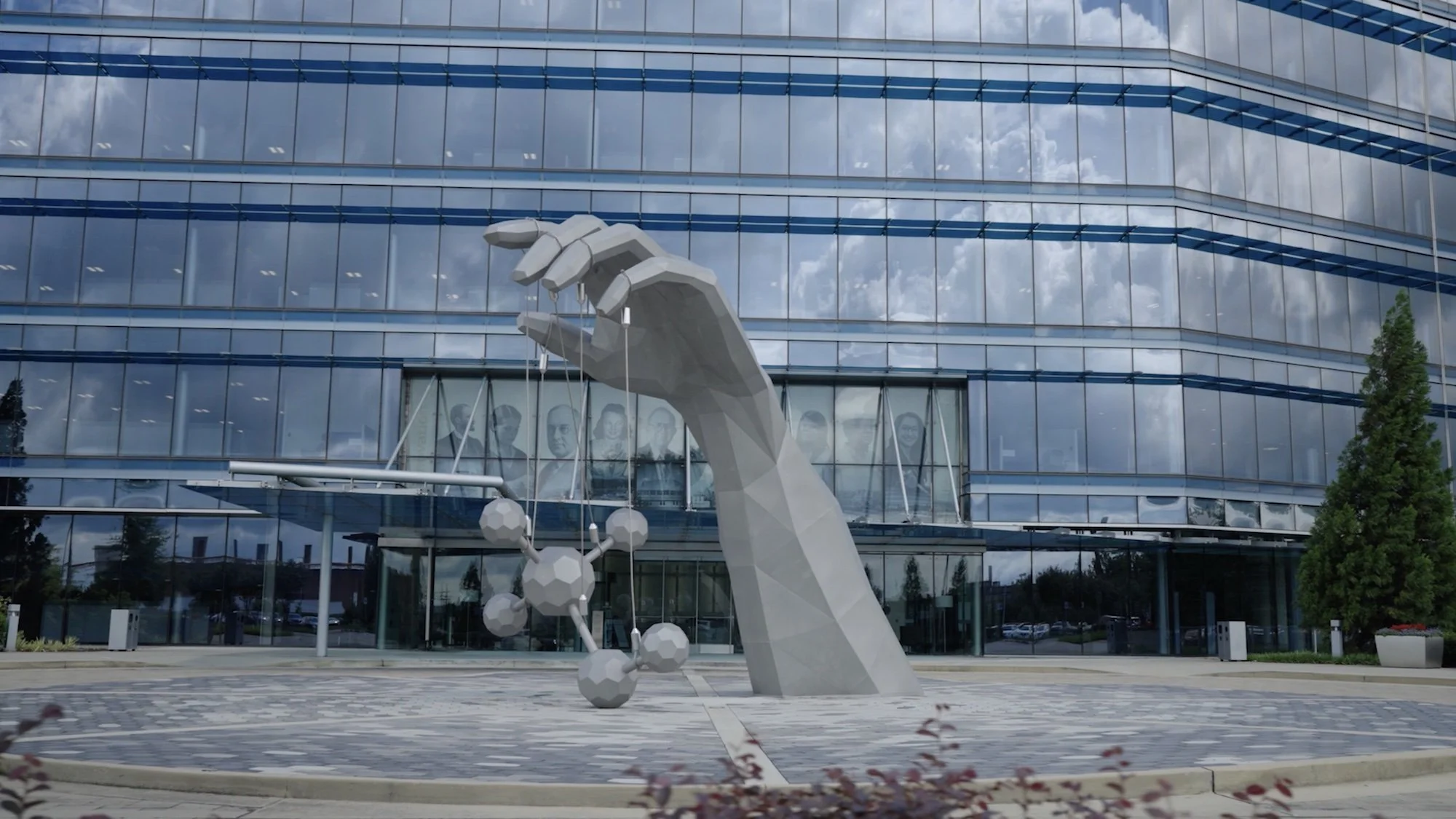 A modern glass building with reflective windows and a monument of a large, stylized hand holding a molecular model outside.