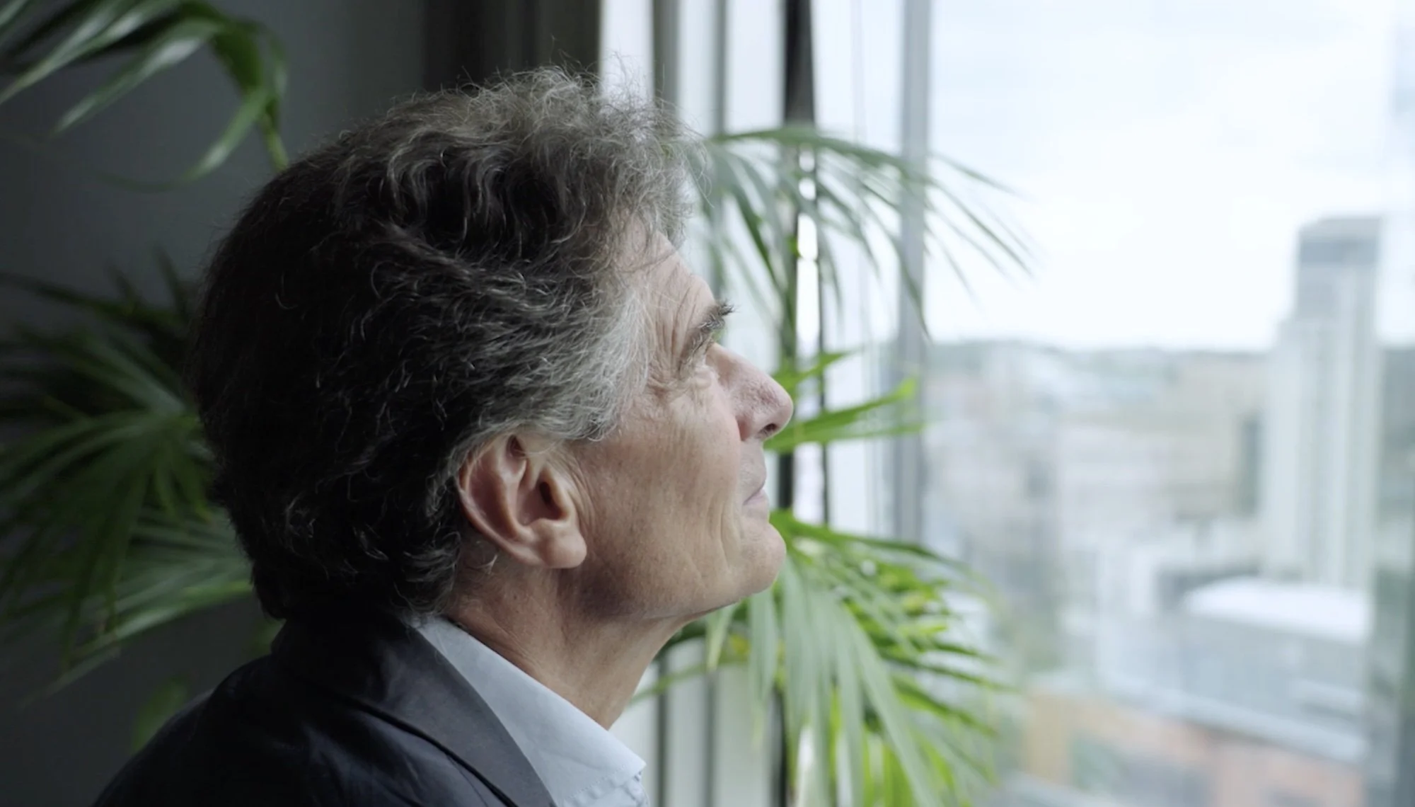 A middle-aged man with gray hair looking out a window with a cityscape in the background, next to green plants.