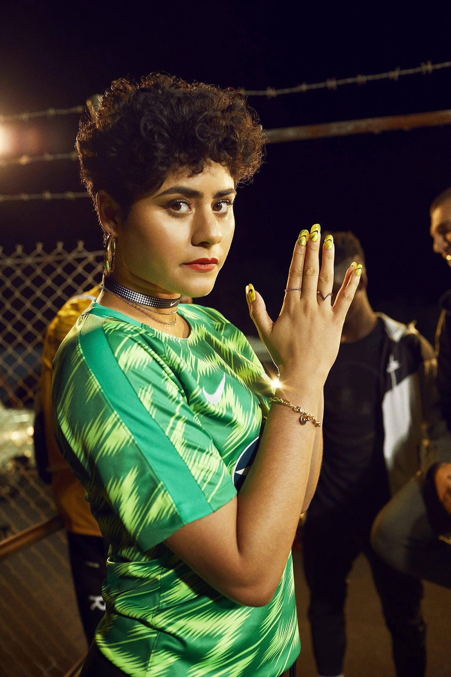 A young woman with short curly hair wearing a green sports jersey, jewelry, and yellow nail polish, raising her hand in a show of strength or solidarity at an outdoor event at night.