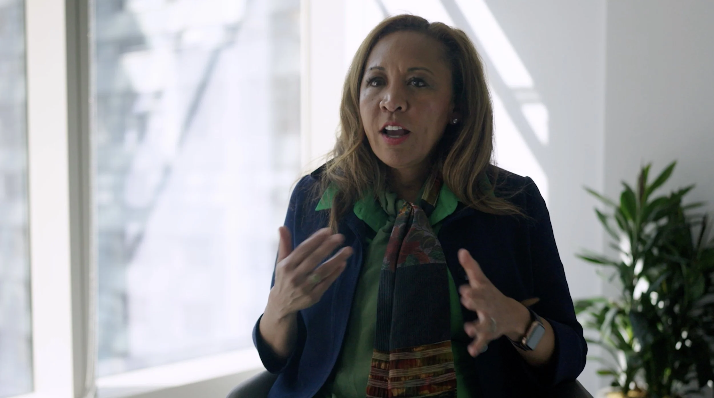 A woman with light brown hair, wearing a dark blazer, green shirt, and patterned scarf, speaking with hand gestures in an office setting near a window with natural light and a plant in the background.