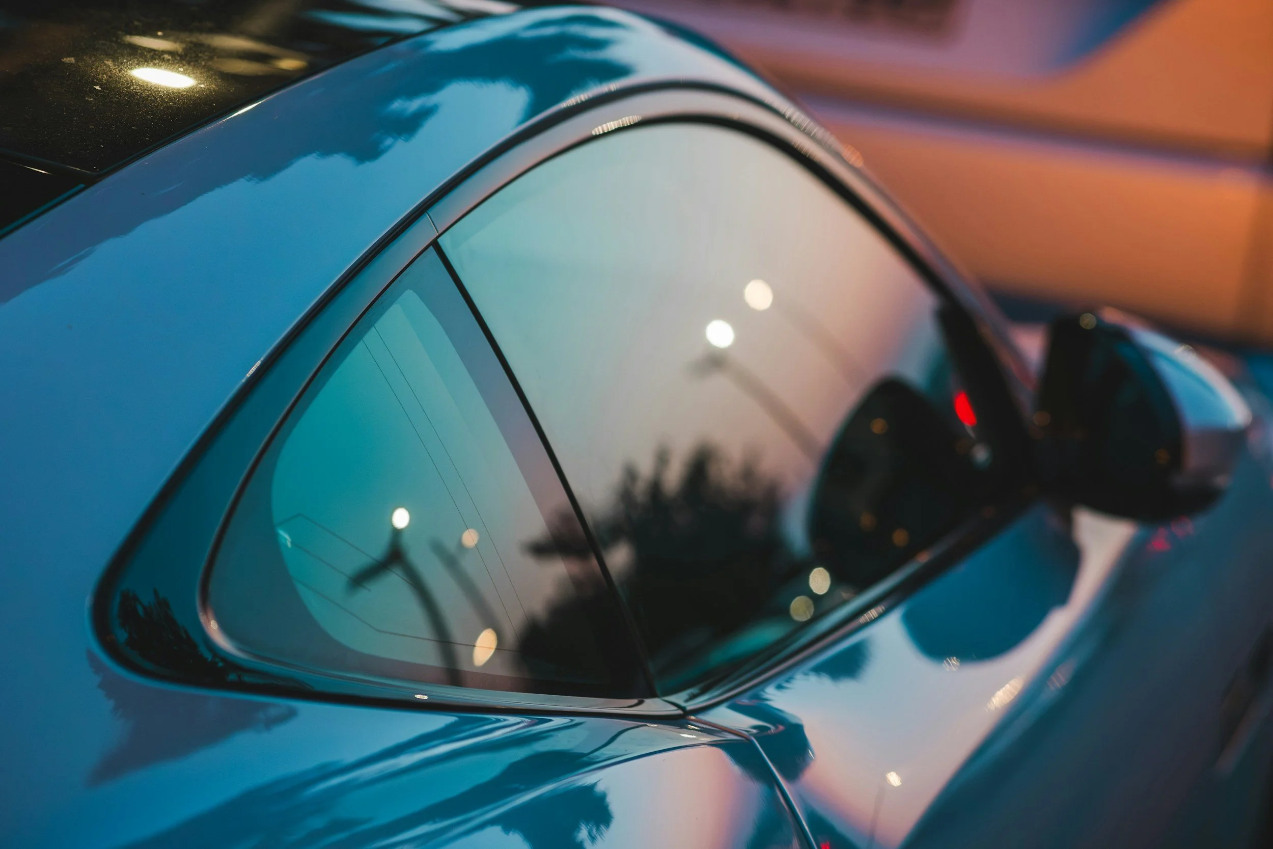Close-up of the rear window and side mirror of a shiny blue car at sunset, with streetlights and trees reflected on the glass.