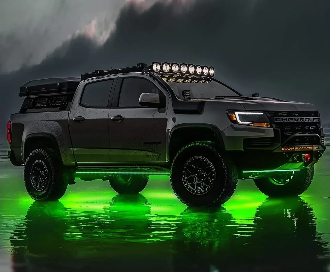 Black Chevrolet Silverado pickup truck with green underglow lights, off-road tires, roof-mounted lights, and a cargo box, parked on wet surface under cloudy sky.