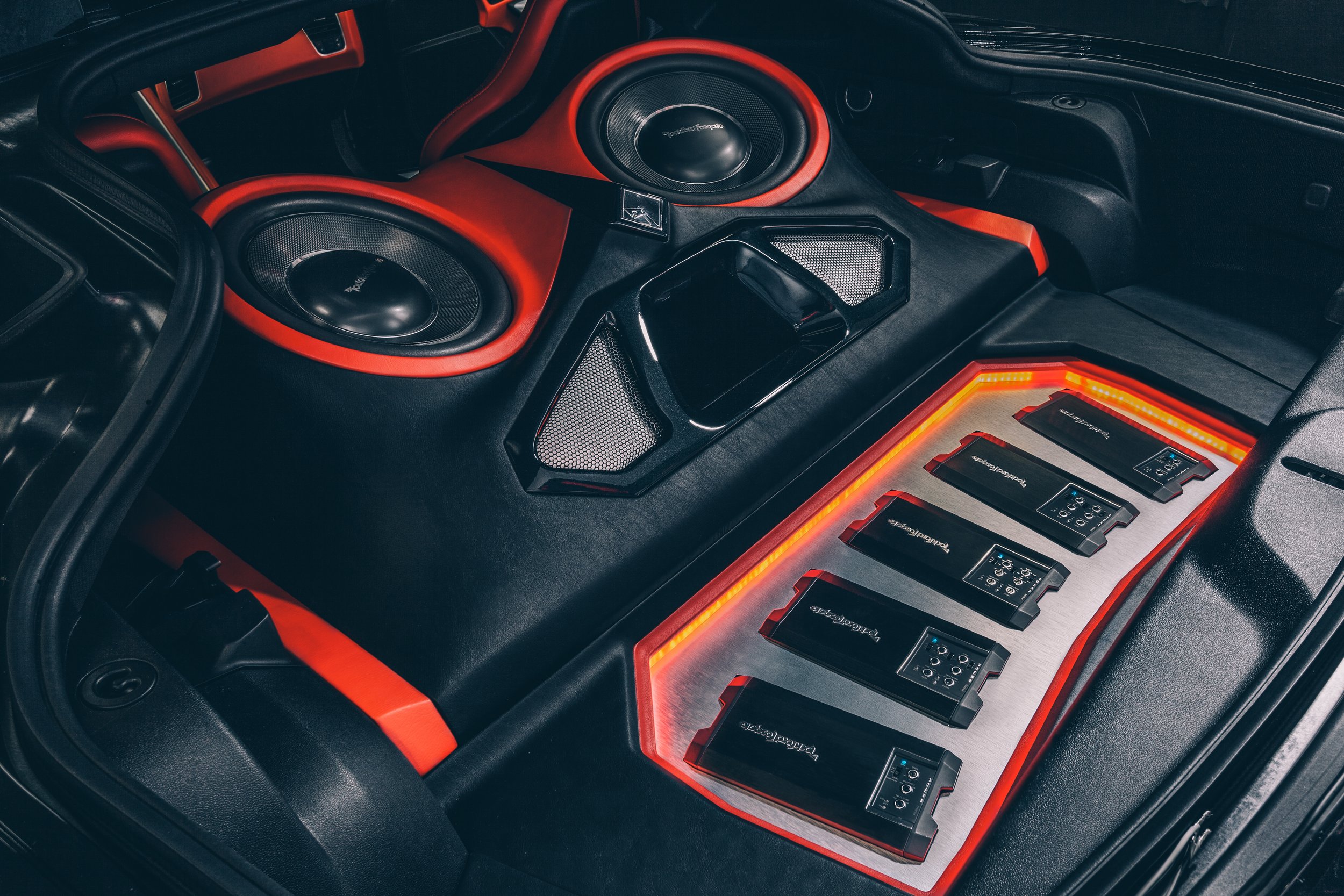 Car interior with custom audio system featuring multiple amplifiers and speakers, illuminated with red and orange lighting.
