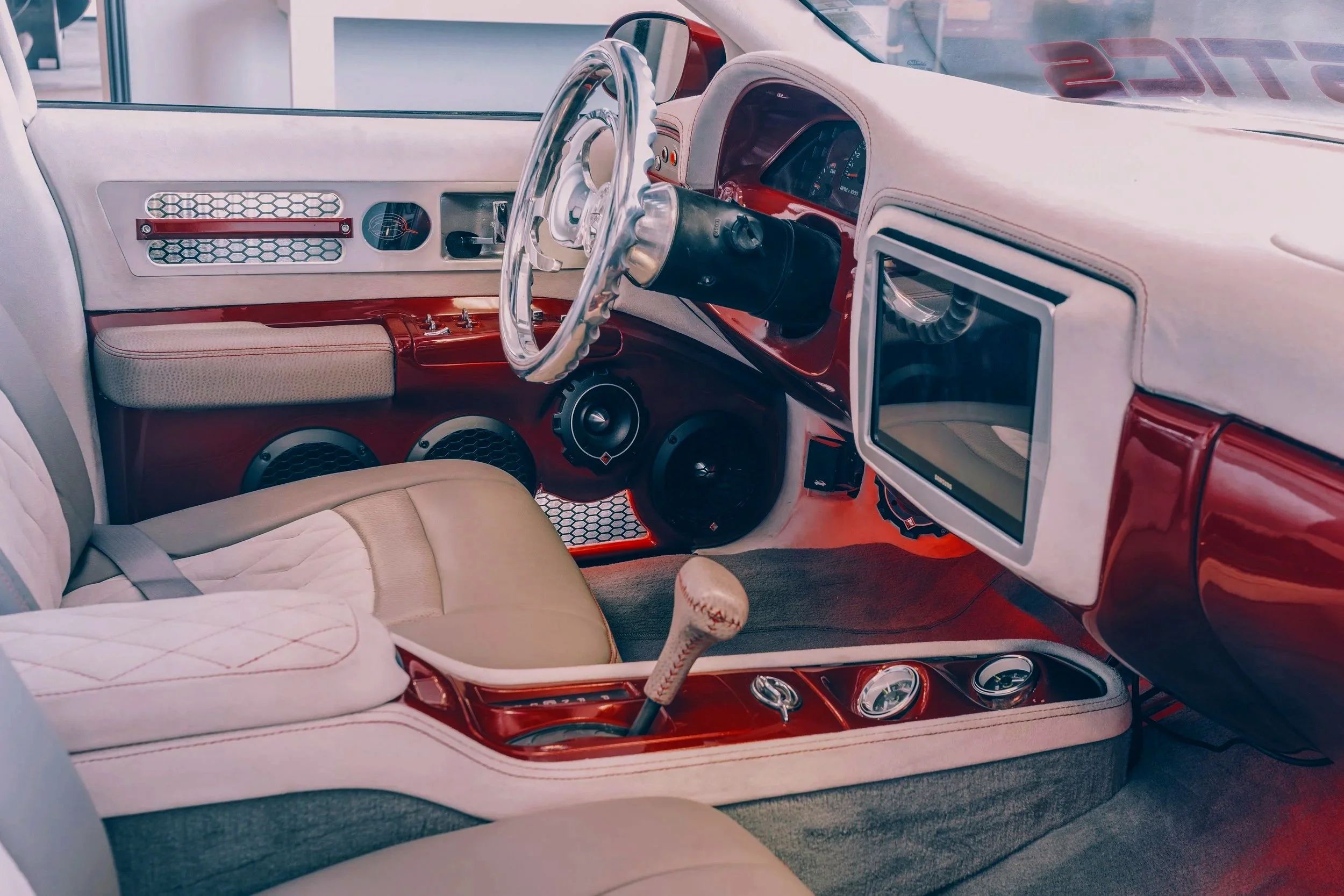 Interior of a vintage car with a beige and red color scheme, featuring a manual gear shift, large speakers, a custom steering wheel, and a TV mounted on the dashboard.