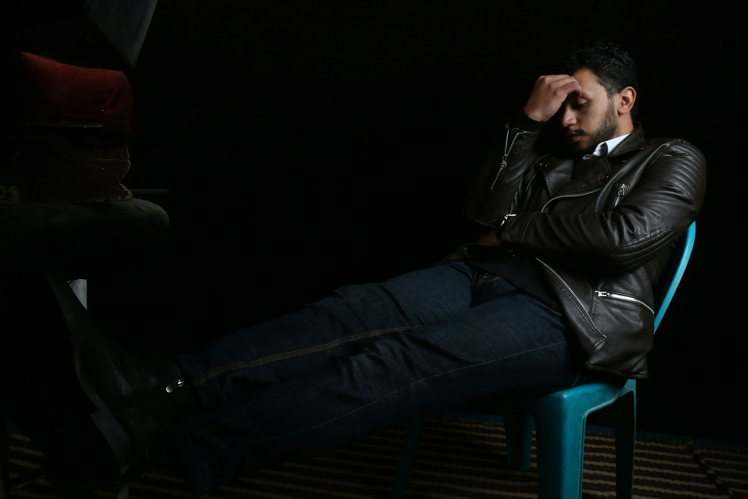 Man sitting on a blue plastic chair, wearing a black leather jacket and dark jeans, with his hand on his forehead, appearing stressed or deep in thought, in a dark room with a black background.