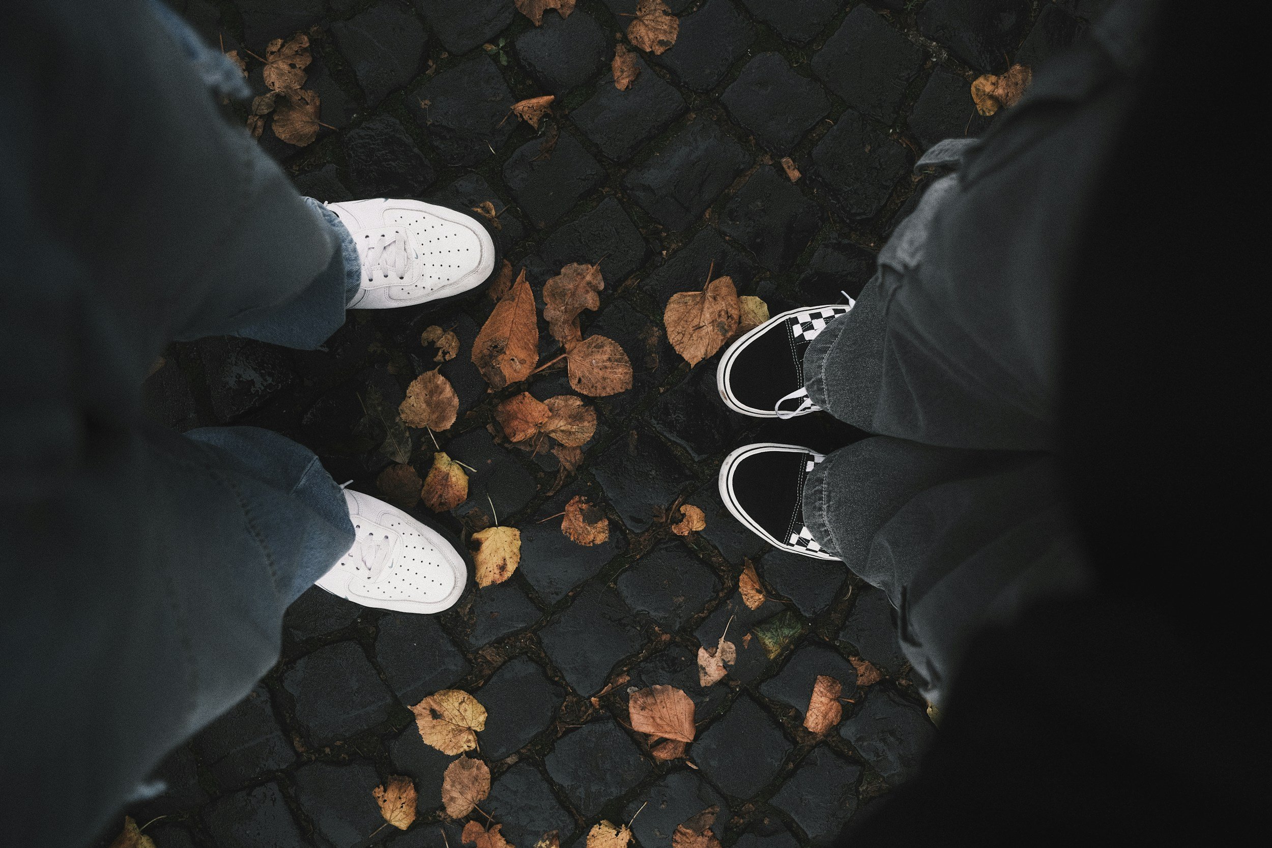 Two people standing on a wet cobblestone street with fallen autumn leaves, wearing sneakers and jeans.