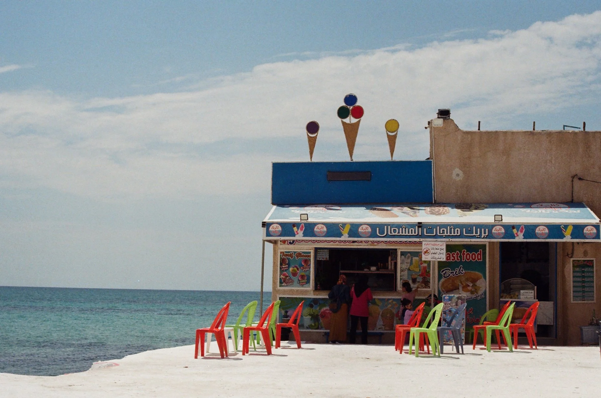 Beachside snack stand with colorful chairs in front, selling ice cream, fast food, and drinks, under a clear sky near the ocean.