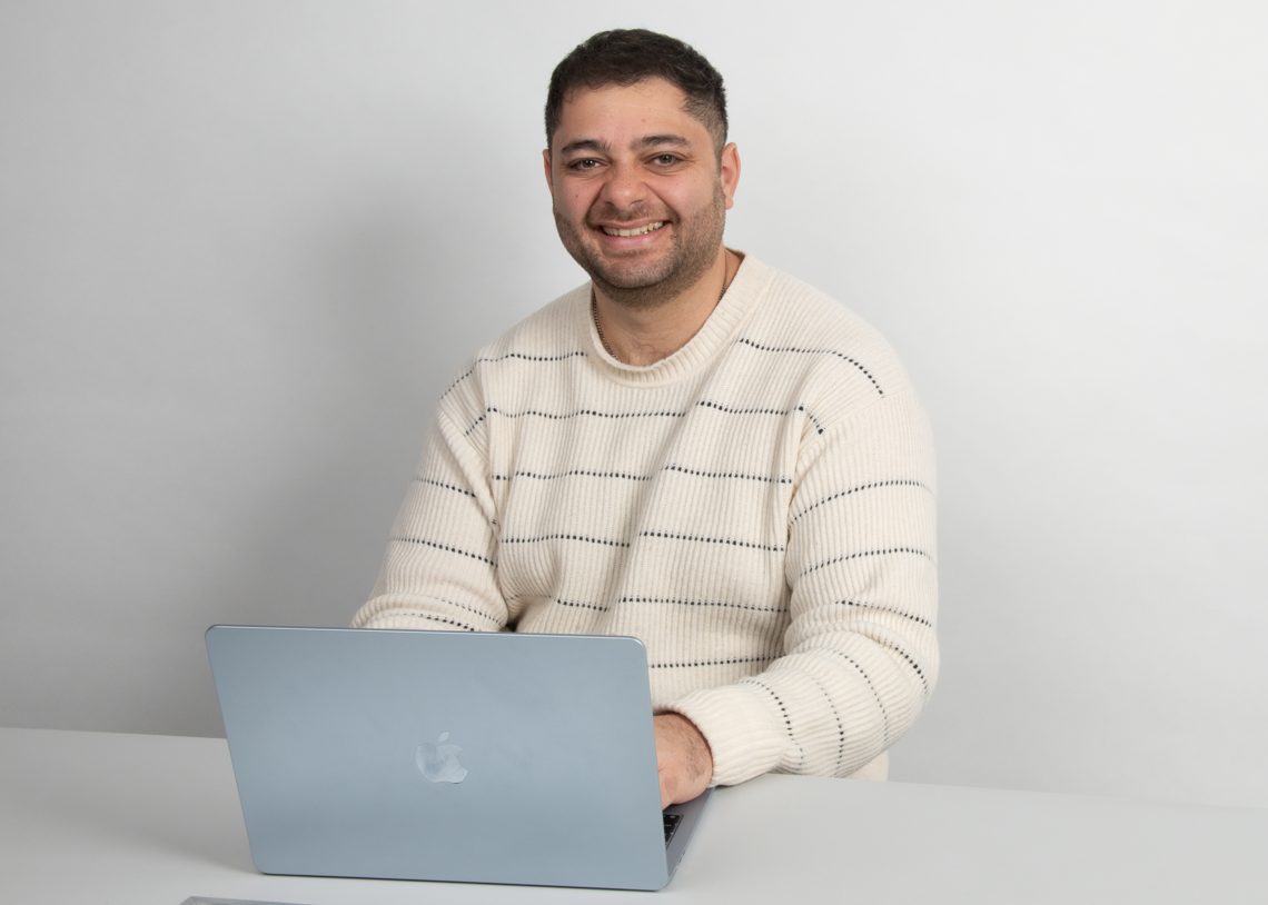 A smiling man sitting at a white table with a light blue MacBook in front of him, against a plain white wall background.