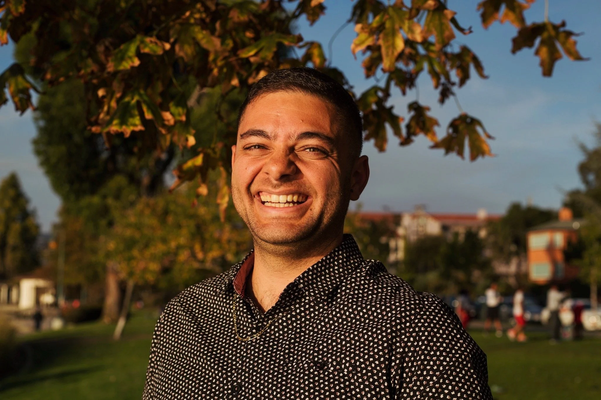 A man smiling outdoors near a tree with autumn-colored leaves, wearing a black and white patterned shirt and a gold chain.