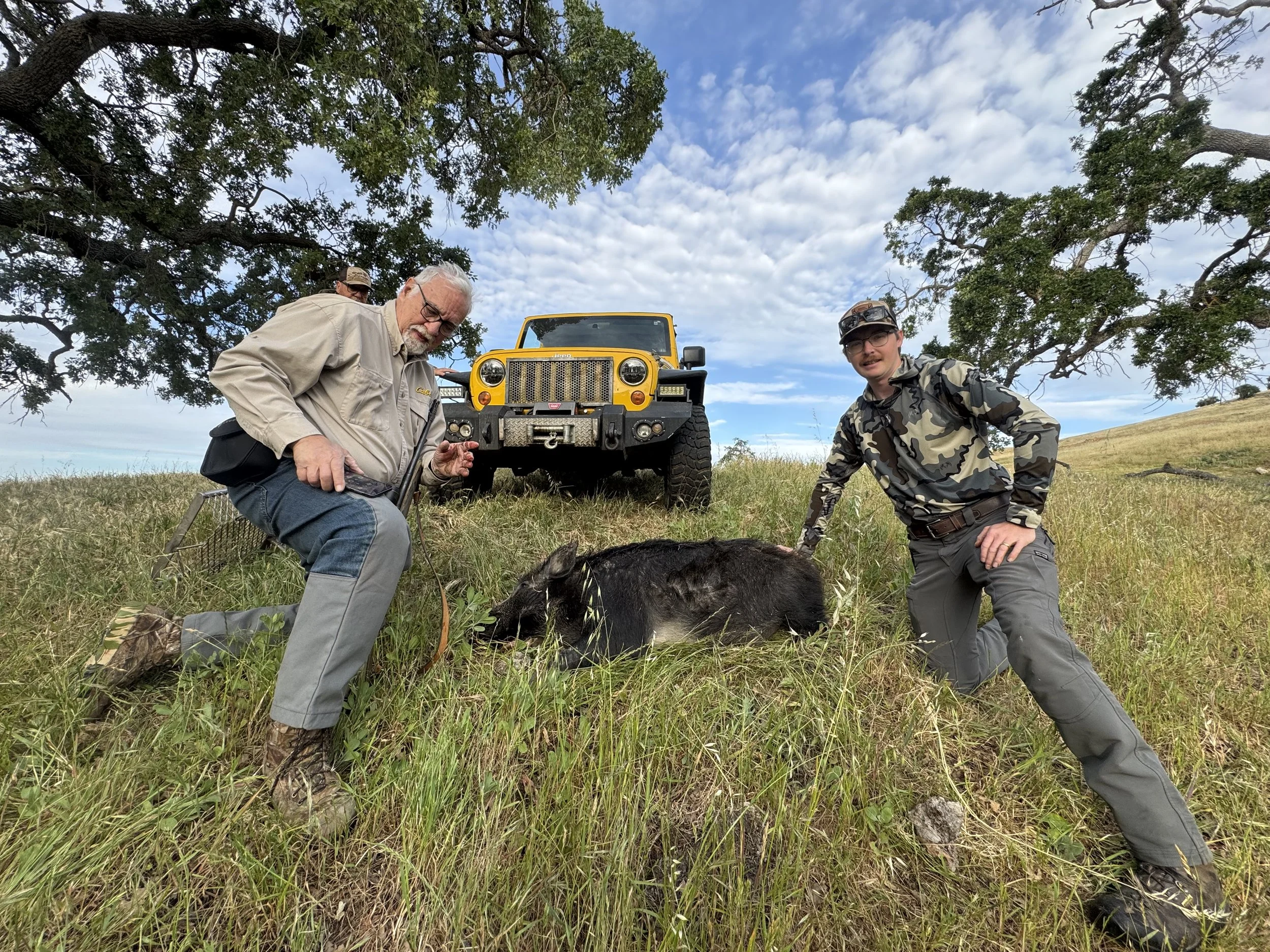 Two men with one older man and one younger man pose outdoors under a large tree with a yellow Jeep in the background. The older man holds a small animal, possibly a rabbit, on the ground. The younger man crouches next to him, wearing camouflage shirt and glasses. The scene appears to be in a grassy field on a partly cloudy day.