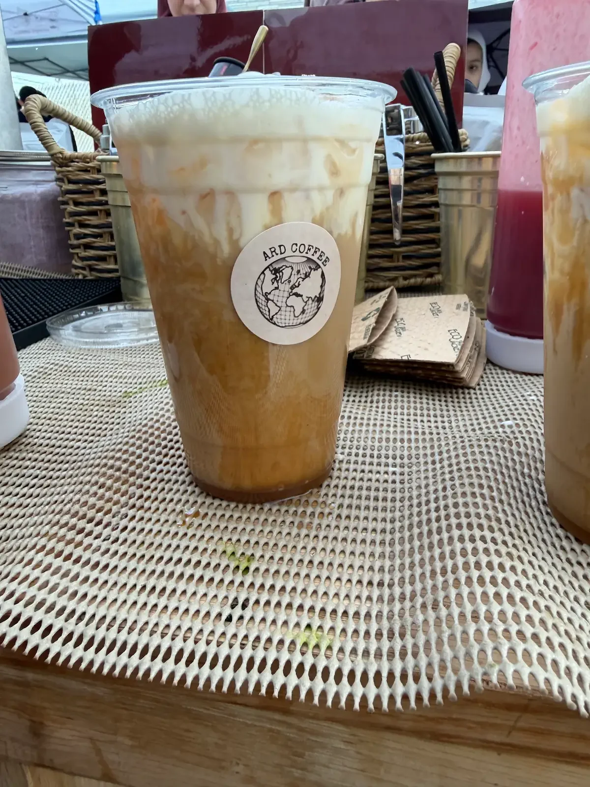A large iced coffee from Ard Coffee with a swirl of cream on top, placed on a woven placemat at an outdoor table.