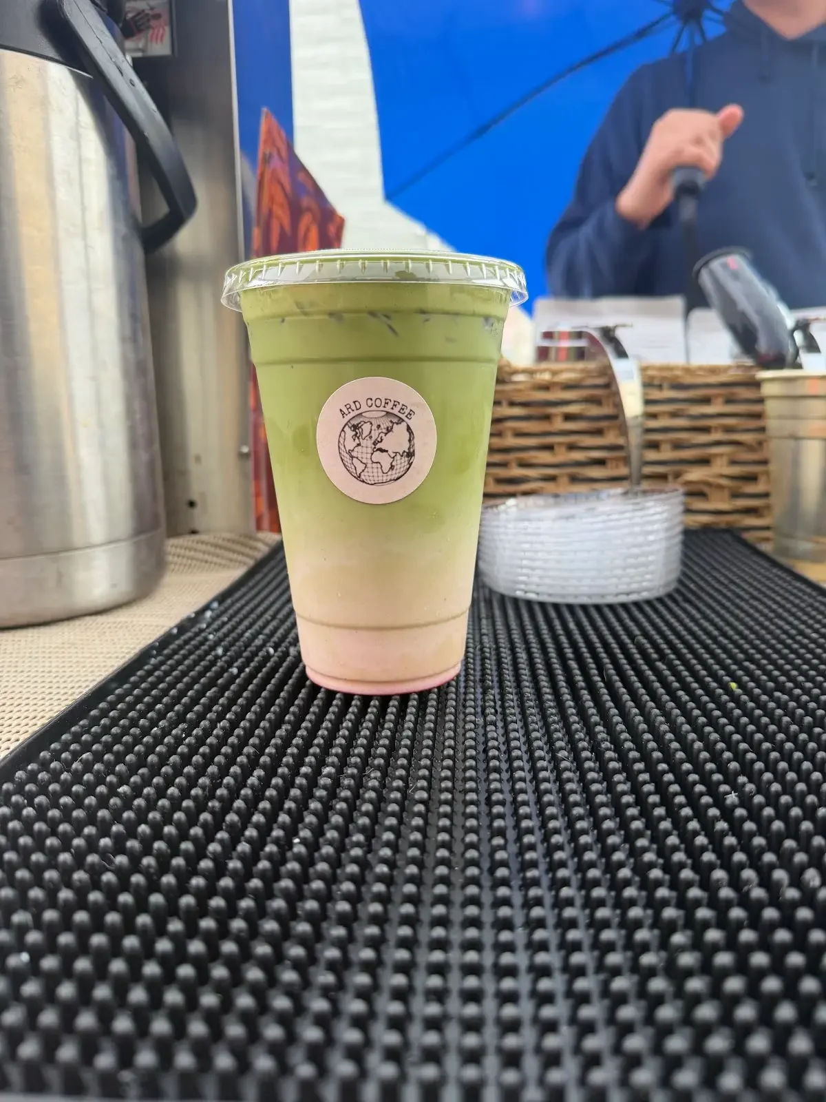 A plastic cup with a layered drink, labeled Ard Coffee, placed on a black rubber mat at a coffee stand. An employee with a blue umbrella and dark jacket is behind the counter, which has wicker baskets and containers.