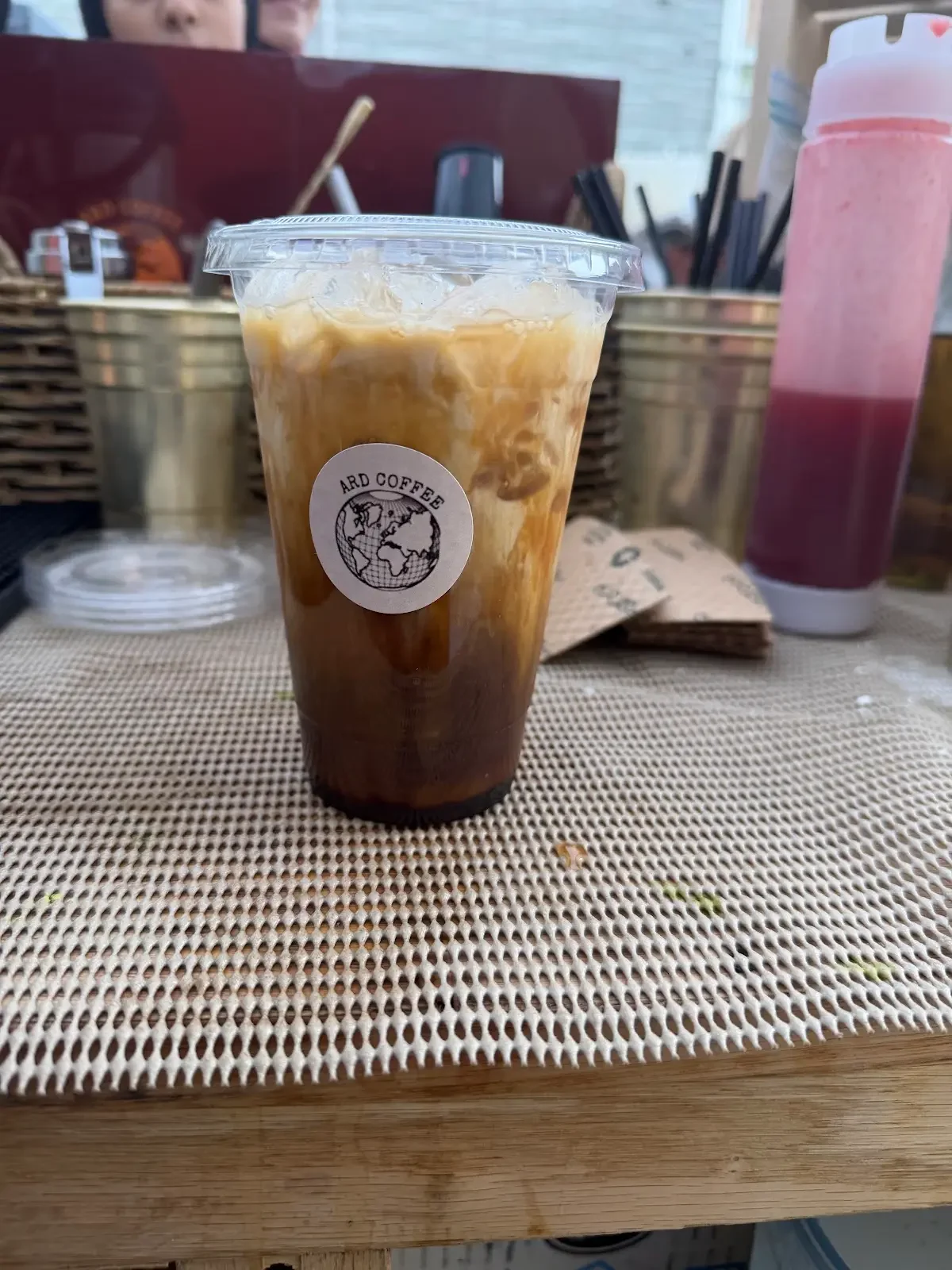 A large plastic cup filled with iced coffee with swirling cream, with a label that says 'ARD COFFEE' and a logo of a globe. There is a pink bottled drink to the right, snacks in the background, and a person partially visible behind the table.